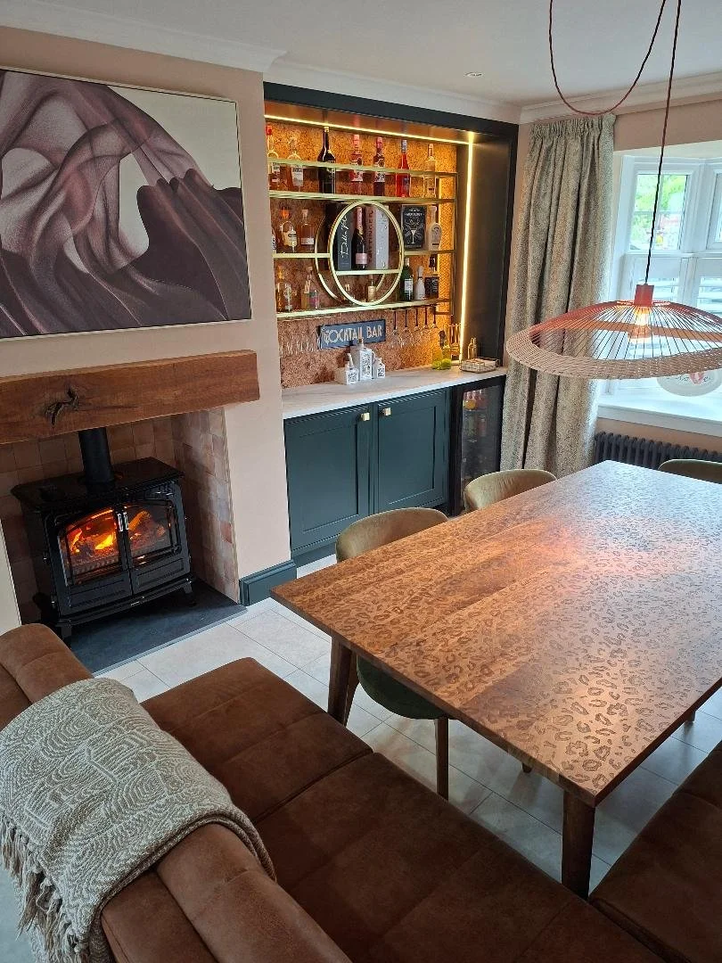 A dining room with leopard print table, cork wall cocktail bar, dark green cabinetry and roaring fire