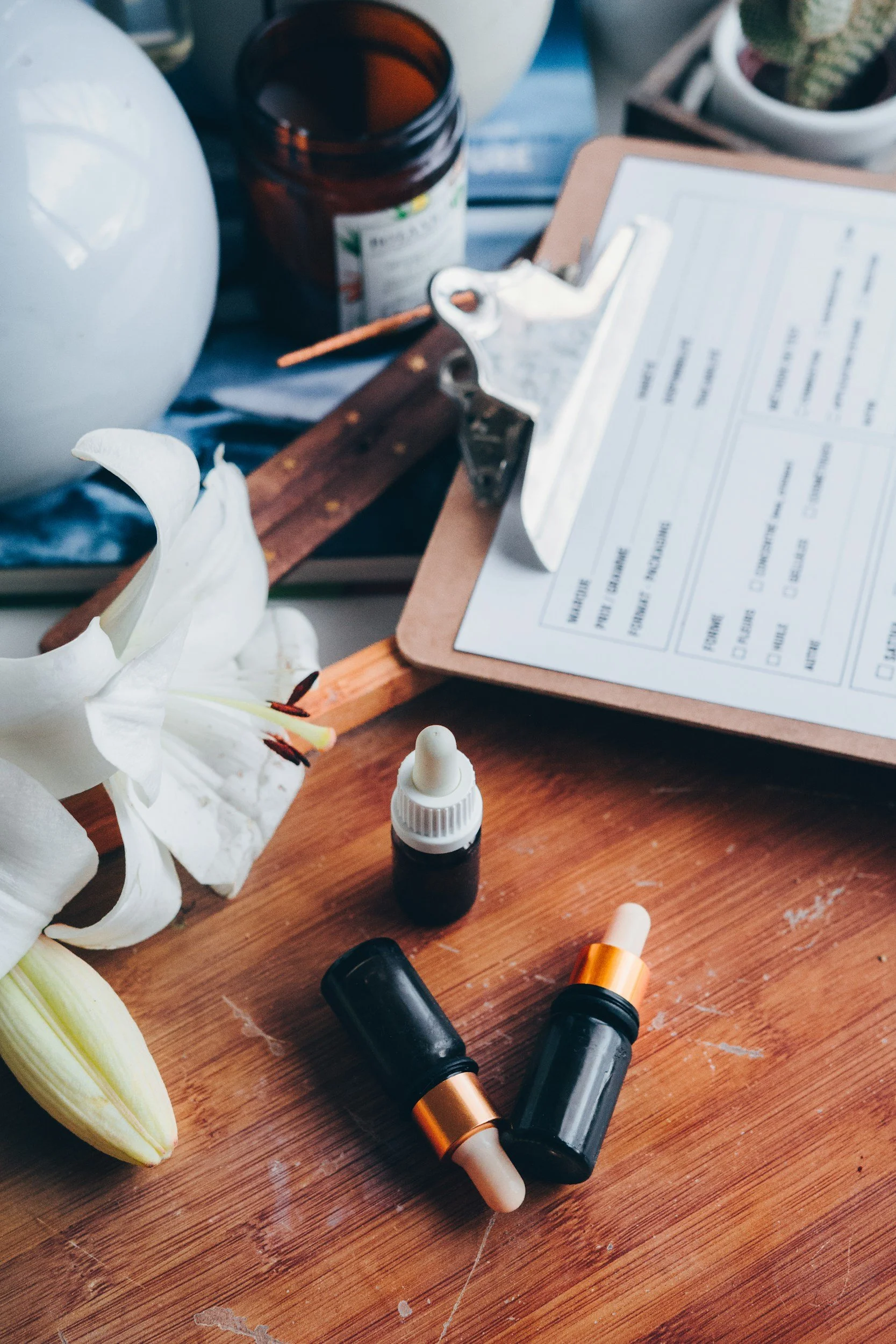 An assortment of items on a wooden surface, including three small bottles with droppers, a white lily flower, an open brown bottle, a clipboard with a paper, and a white ceramic pot with a cactus.