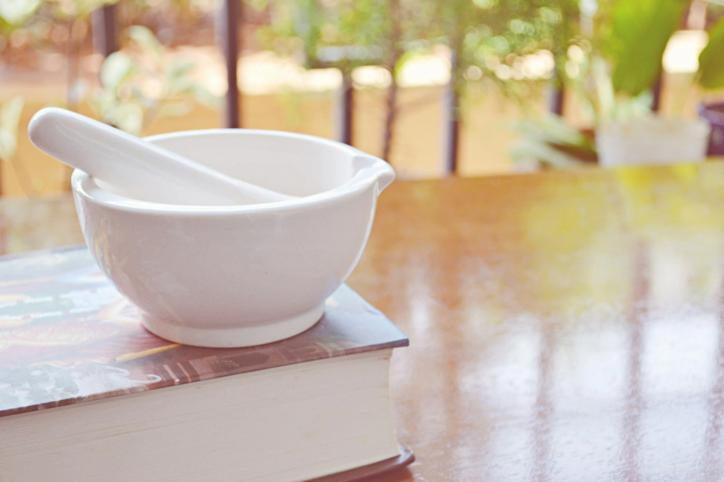 White ceramic mortar and pestle on a wooden table with a blurred background of green foliage and sunlight.