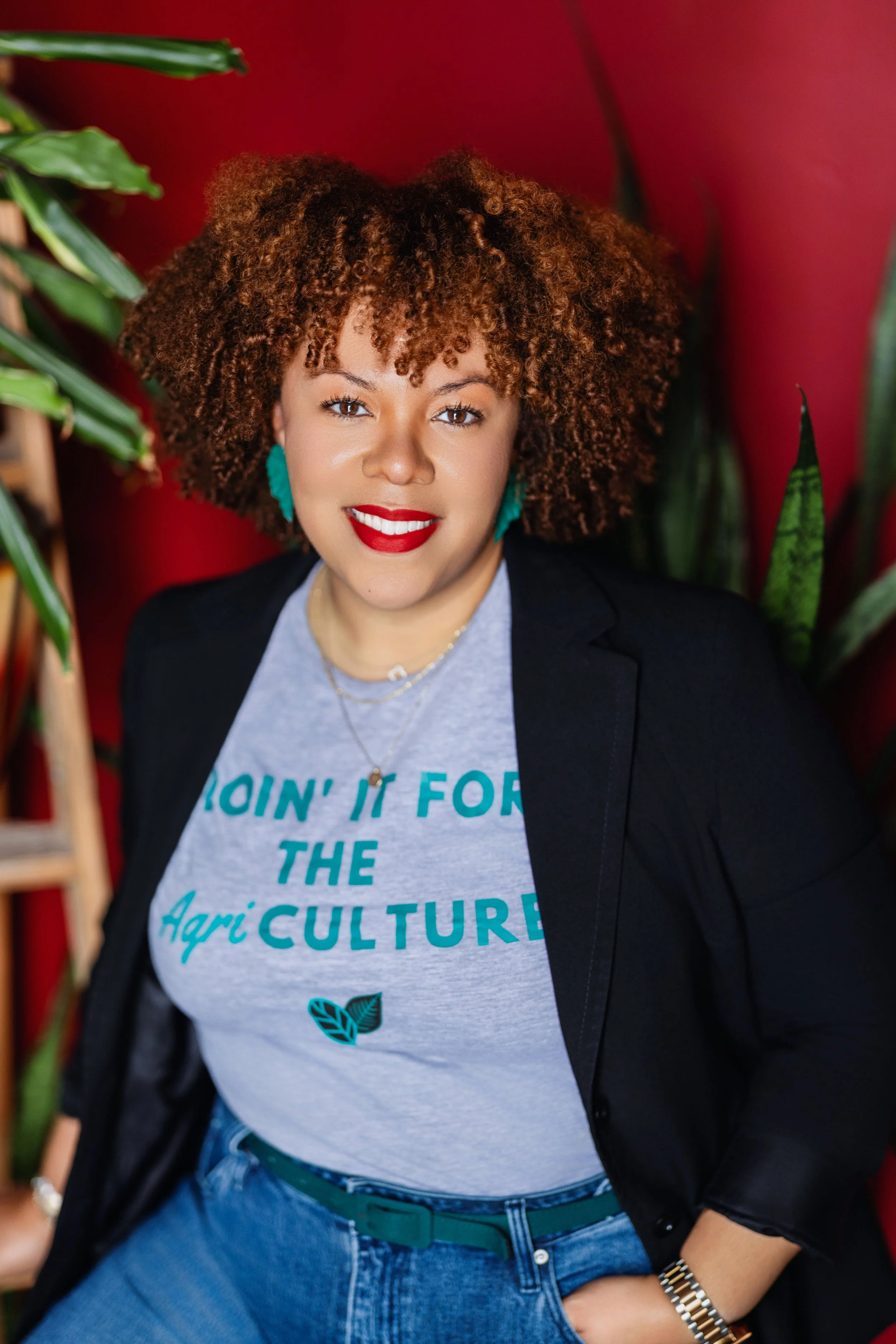 Close-up of a woman with curly, reddish-brown hair, red lipstick, teal earrings, wearing a graphic t-shirt with text about agriculture, layered necklaces, and a black blazer, standing against a red background surrounded by green plants.