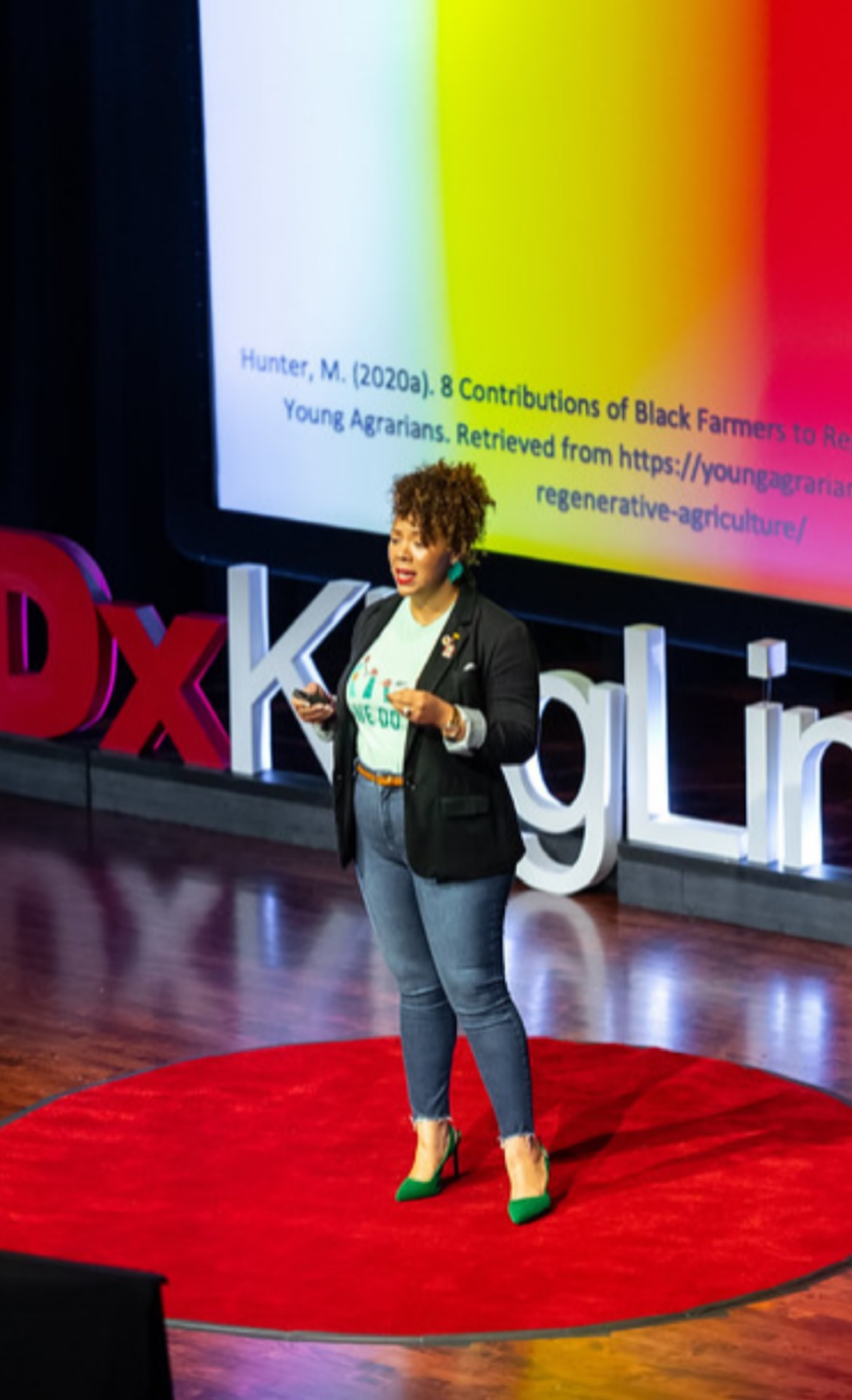 A woman on a red circular stage giving a presentation at a TEDx event, with large 3D letters spelling TEDx and a screen behind her displaying a scientific paper citation.