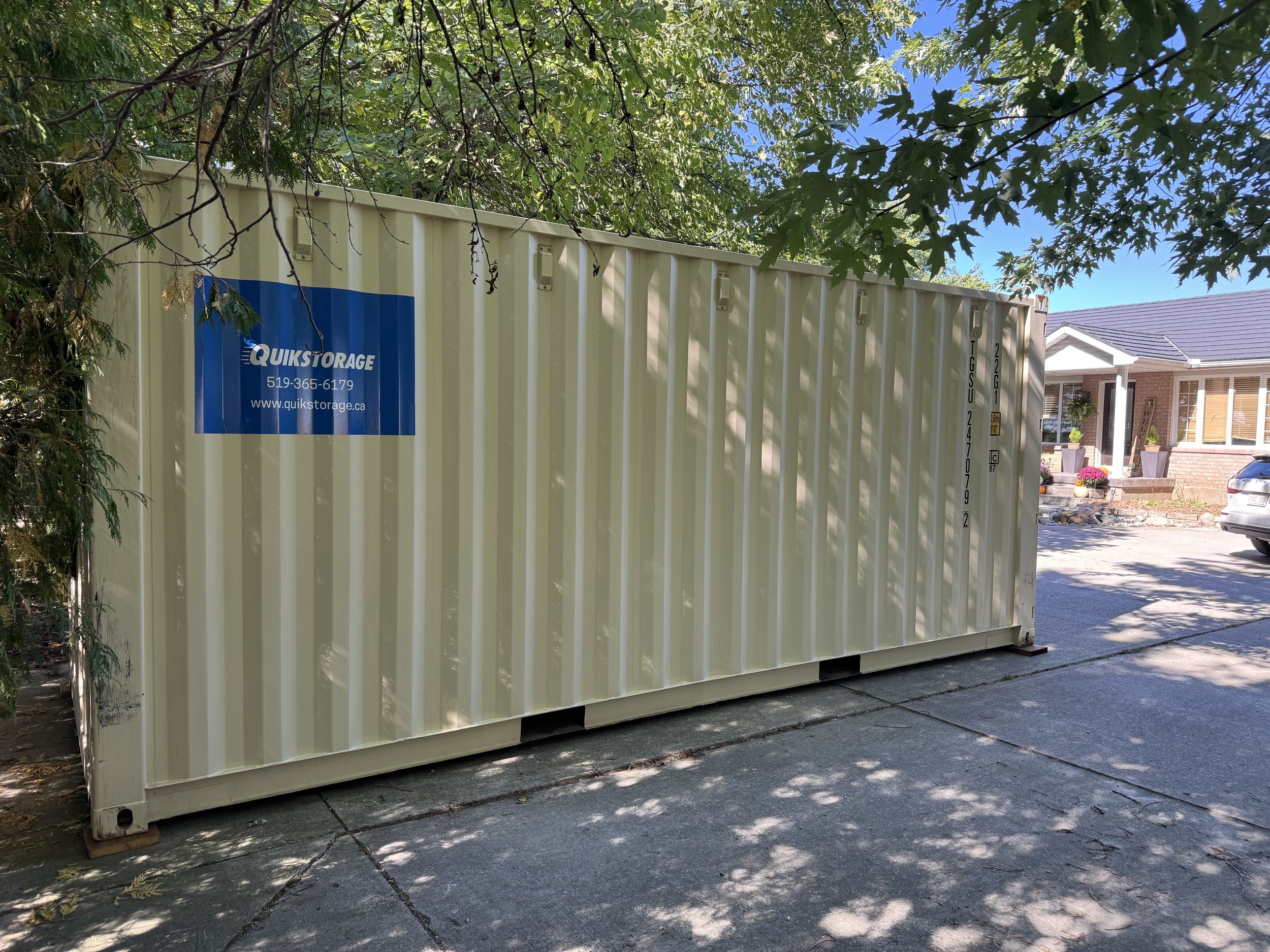 A yellow storage container with a blue Quik Storage logo, situated outdoors among trees and near a residential house, under a partly cloudy sky.