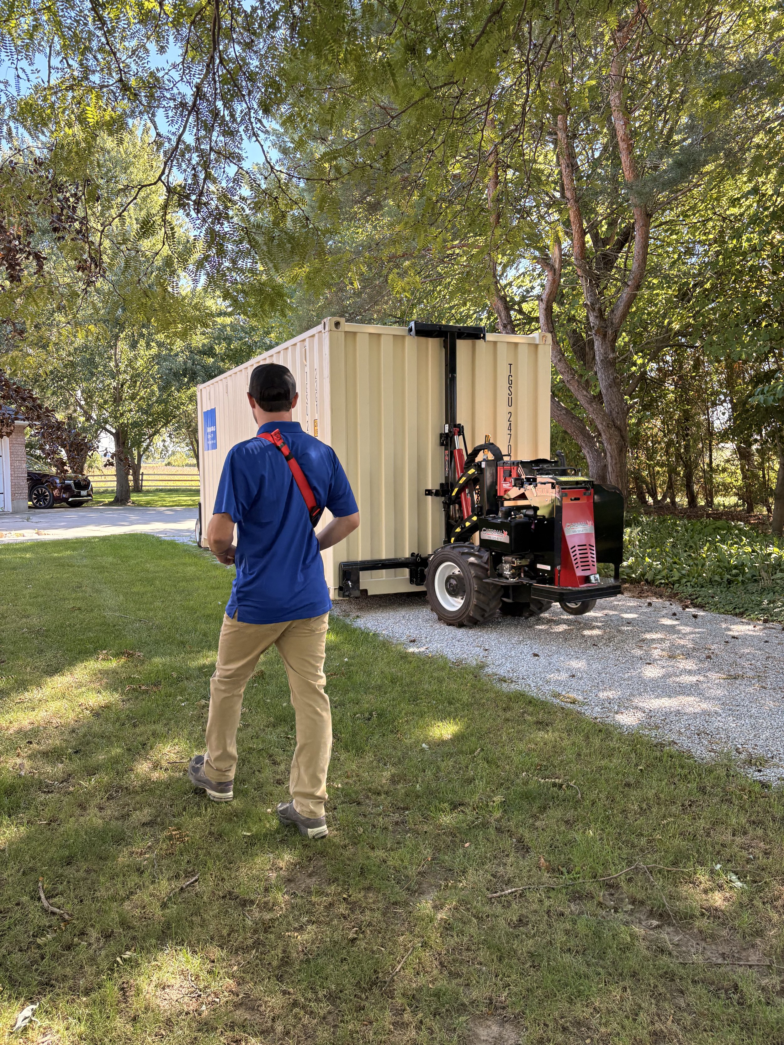 A man in a blue shirt and beige pants walking on grass in front of a large tan shipping container, with a red and black machine nearby, under leafy trees on a sunny day.
