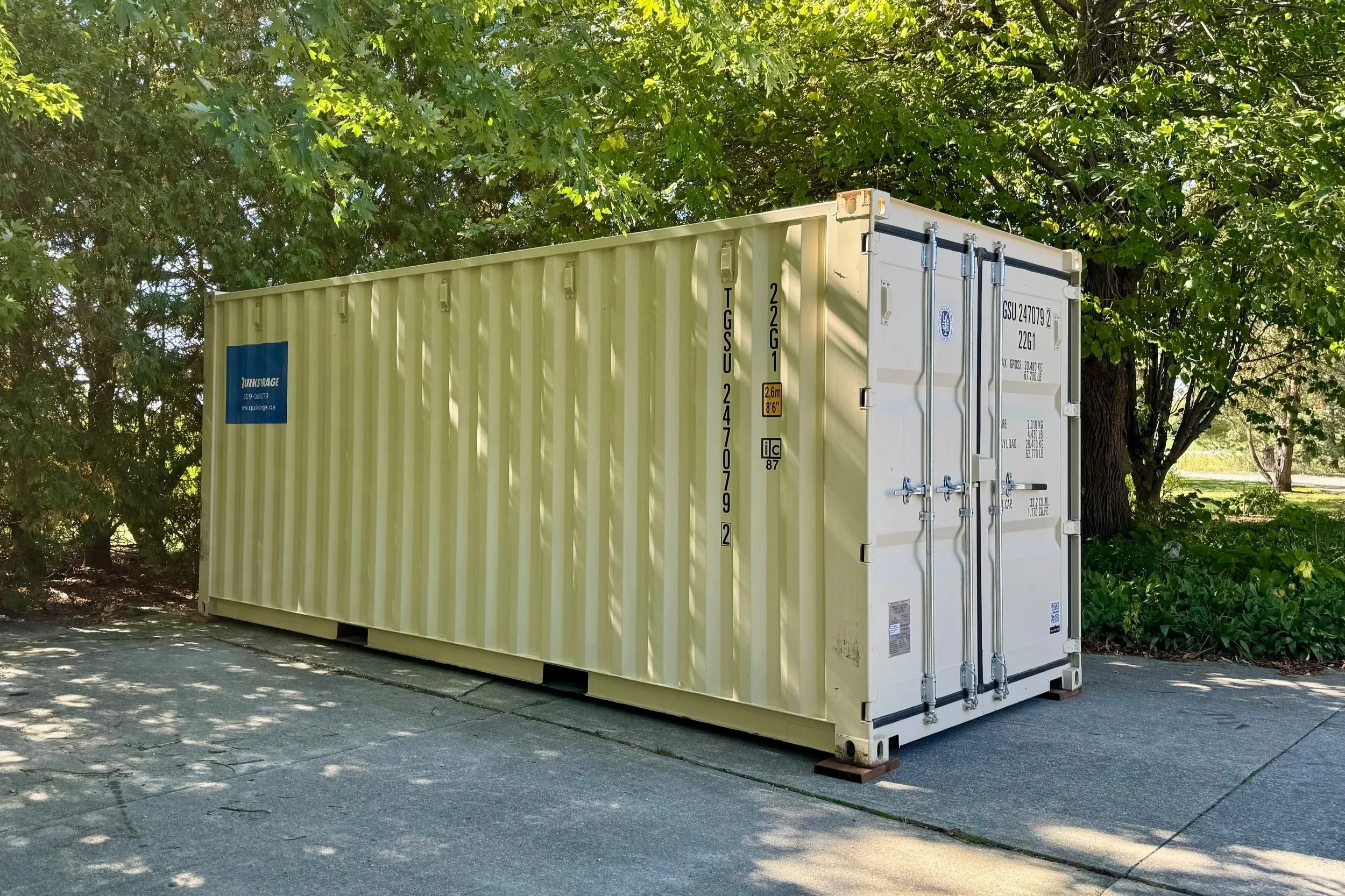 A yellow shipping container with a blue logo and text, situated outdoors on a concrete surface, surrounded by green trees and foliage.
