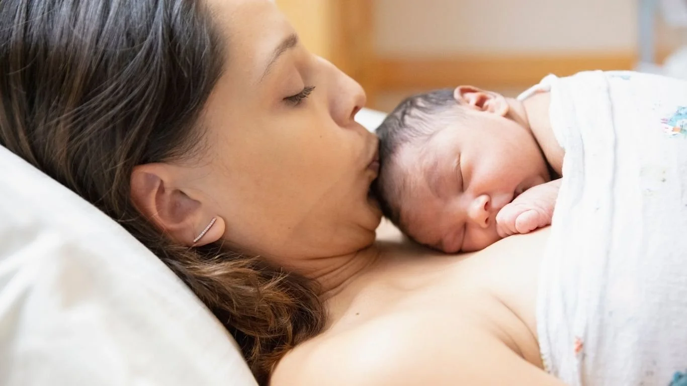 A woman with long brown hair and earrings lying down with her eyes closed, holding a sleeping newborn baby with dark hair close to her chest.