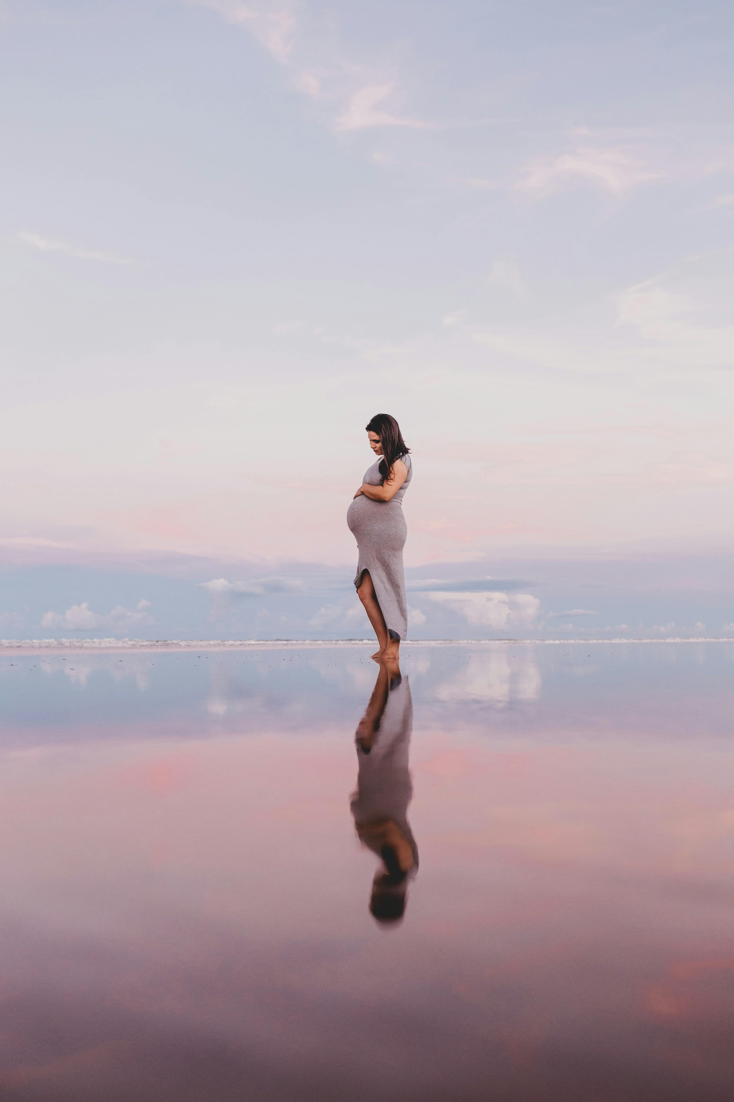 A pregnant woman in a long gray dress standing on a reflective surface, with a pastel-colored sky in the background.