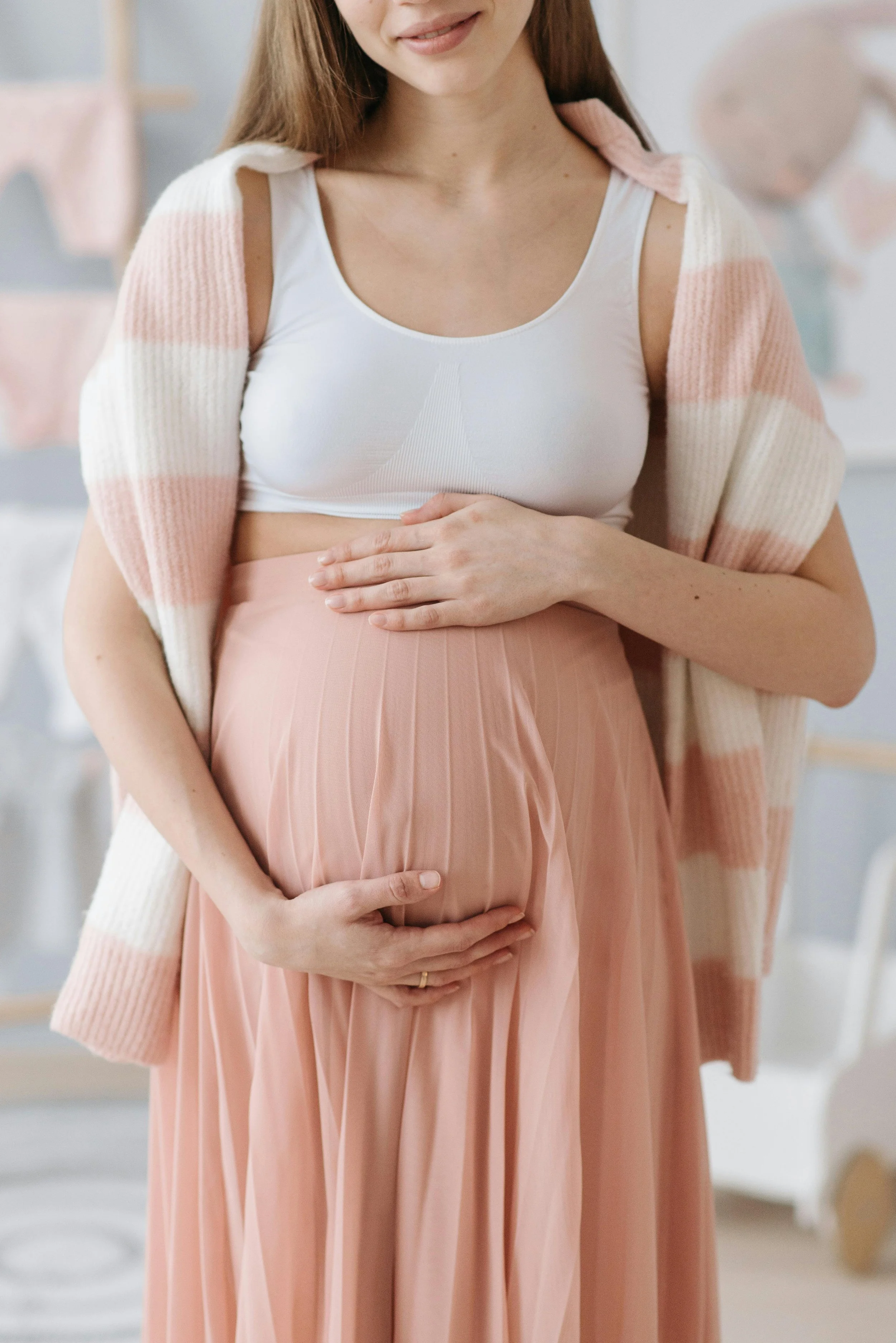 A pregnant woman is gently holding her belly with both hands while standing indoors in front of a soft background. She is wearing a white crop top, a pink pleated skirt, and a cardigan draped over her shoulders.