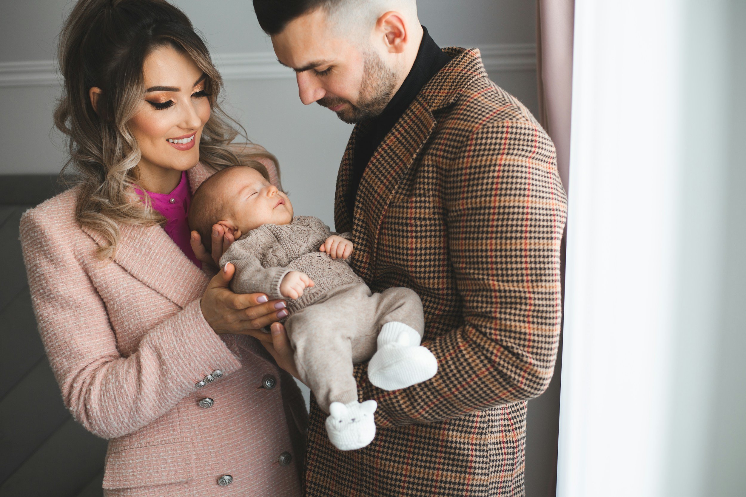 A woman and man are holding a sleeping baby between them, smiling and looking at the child lovingly. They are indoors near a window with curtains.