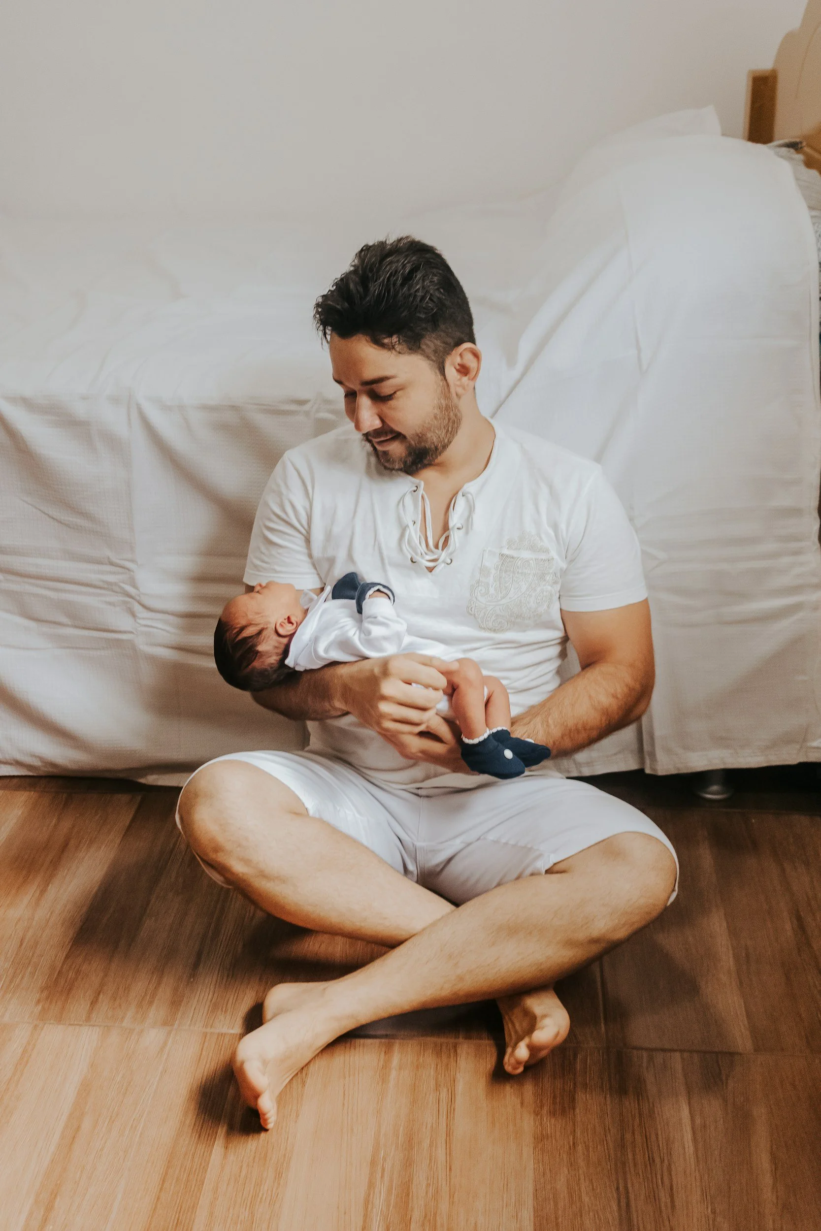 A man with dark hair and a beard sitting cross-legged on wooden floor, holding a small baby boy in his arms, both looking fondly at each other in a room with a bed and white bedding in the background.