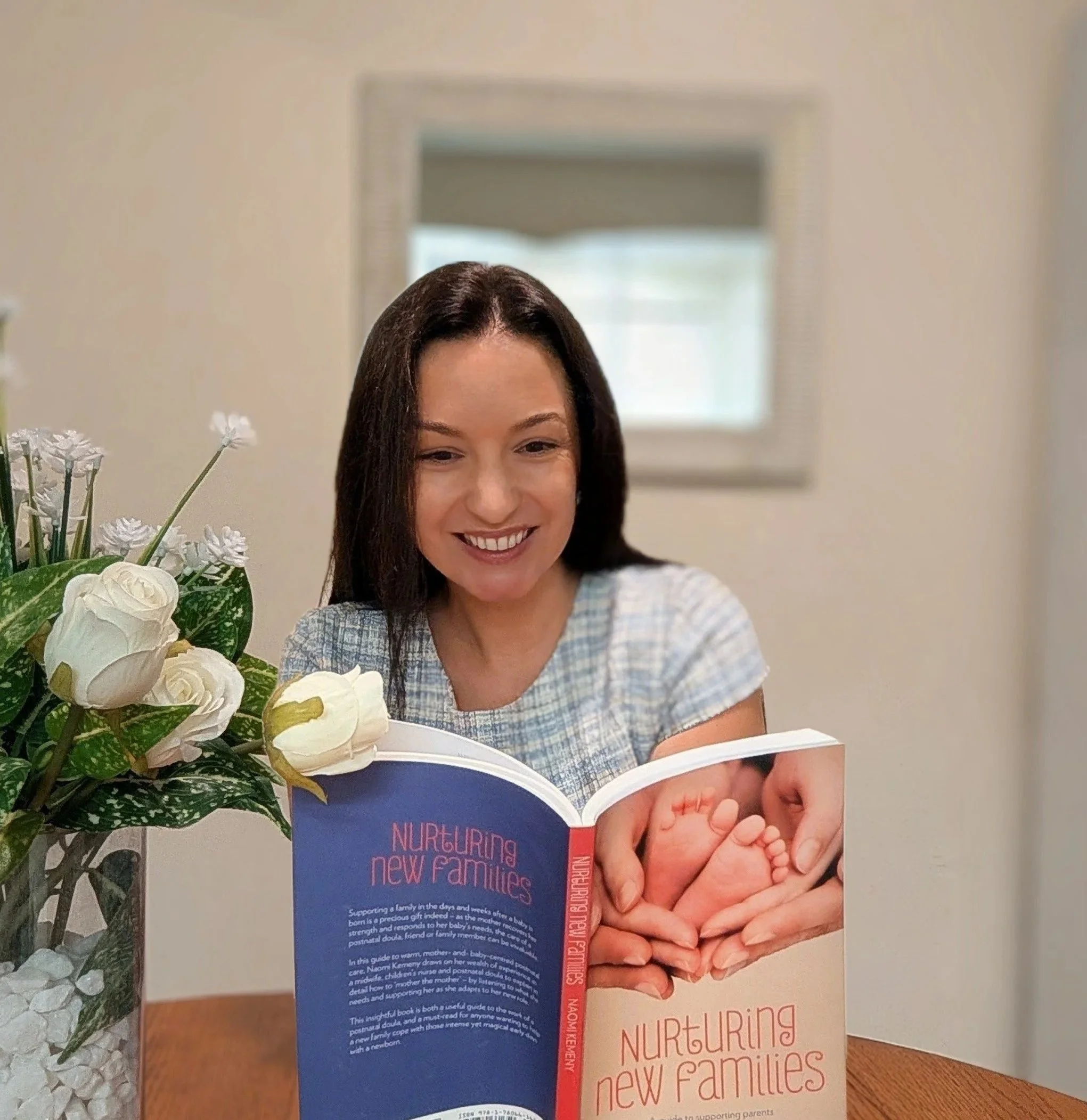 A woman with dark brown hair smiling while holding a baby. There are white flowers and a book titled 'Nurturing New Families' in the foreground, and a mirror reflecting the room in the background.