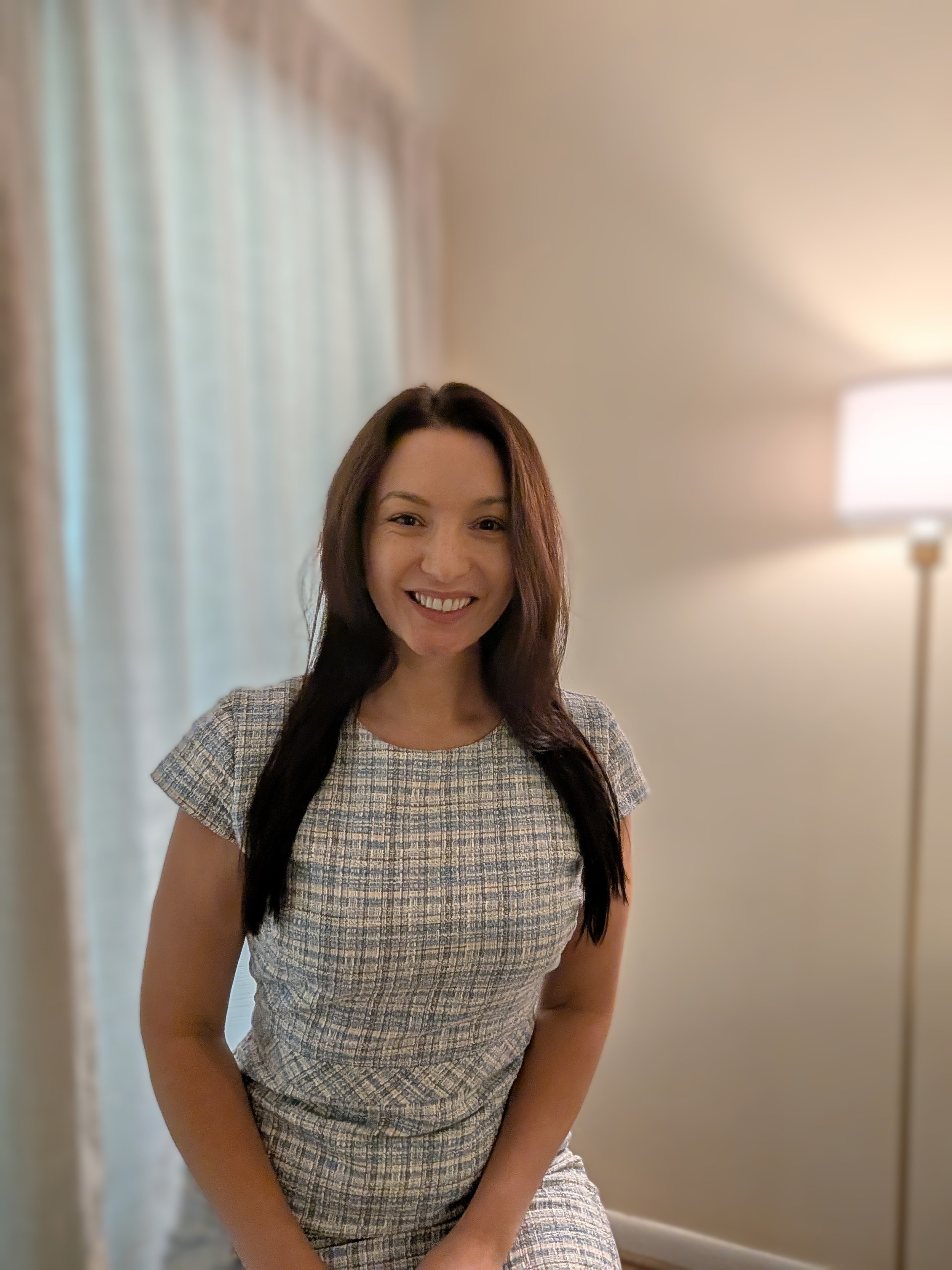 A woman with long dark hair wearing a grey checkered dress, smiling, standing indoors near a wall with a lamp in the background.