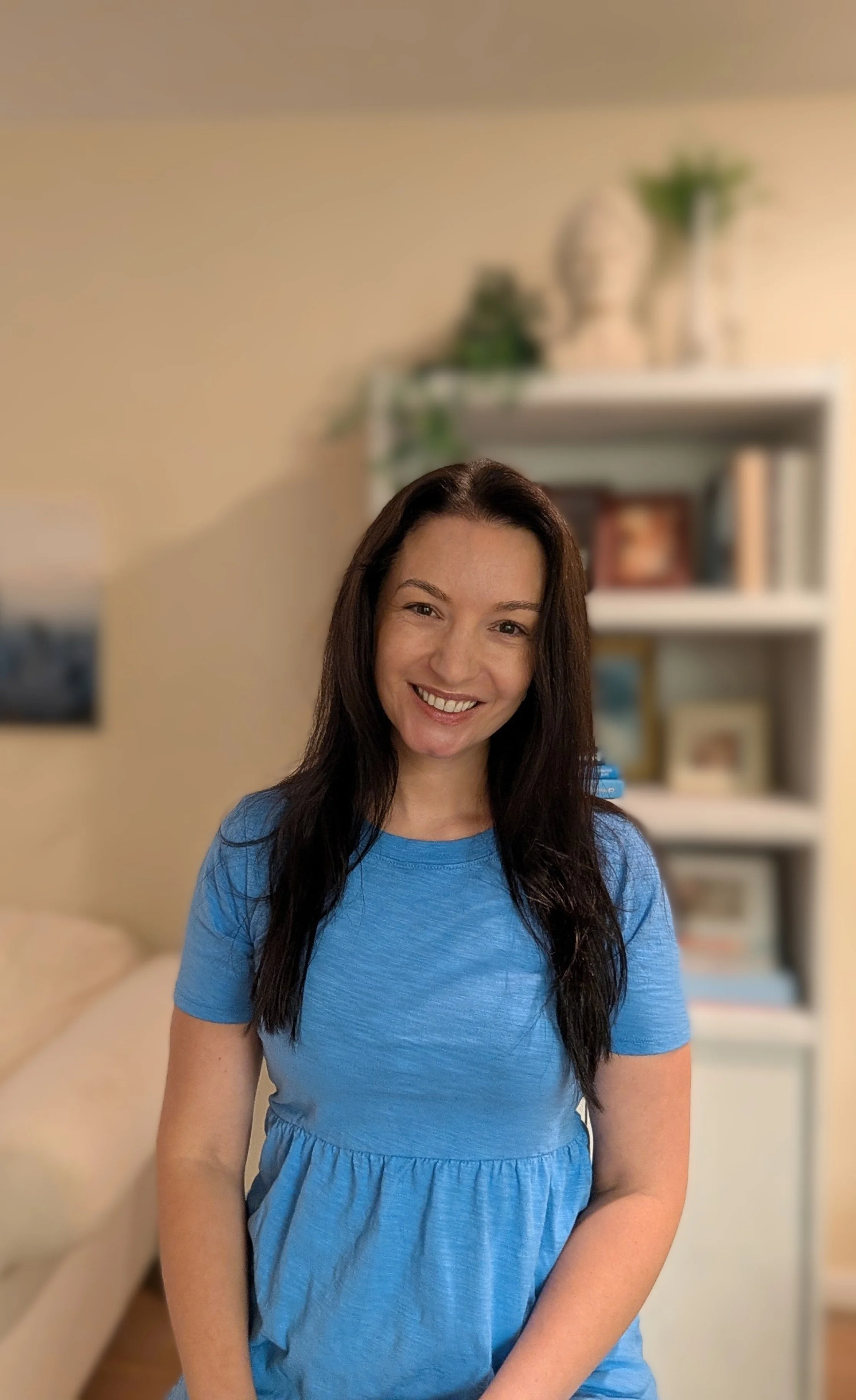 A woman with long dark hair smiling, wearing a blue dress, standing indoors with a beige wall and bookshelf in the background.