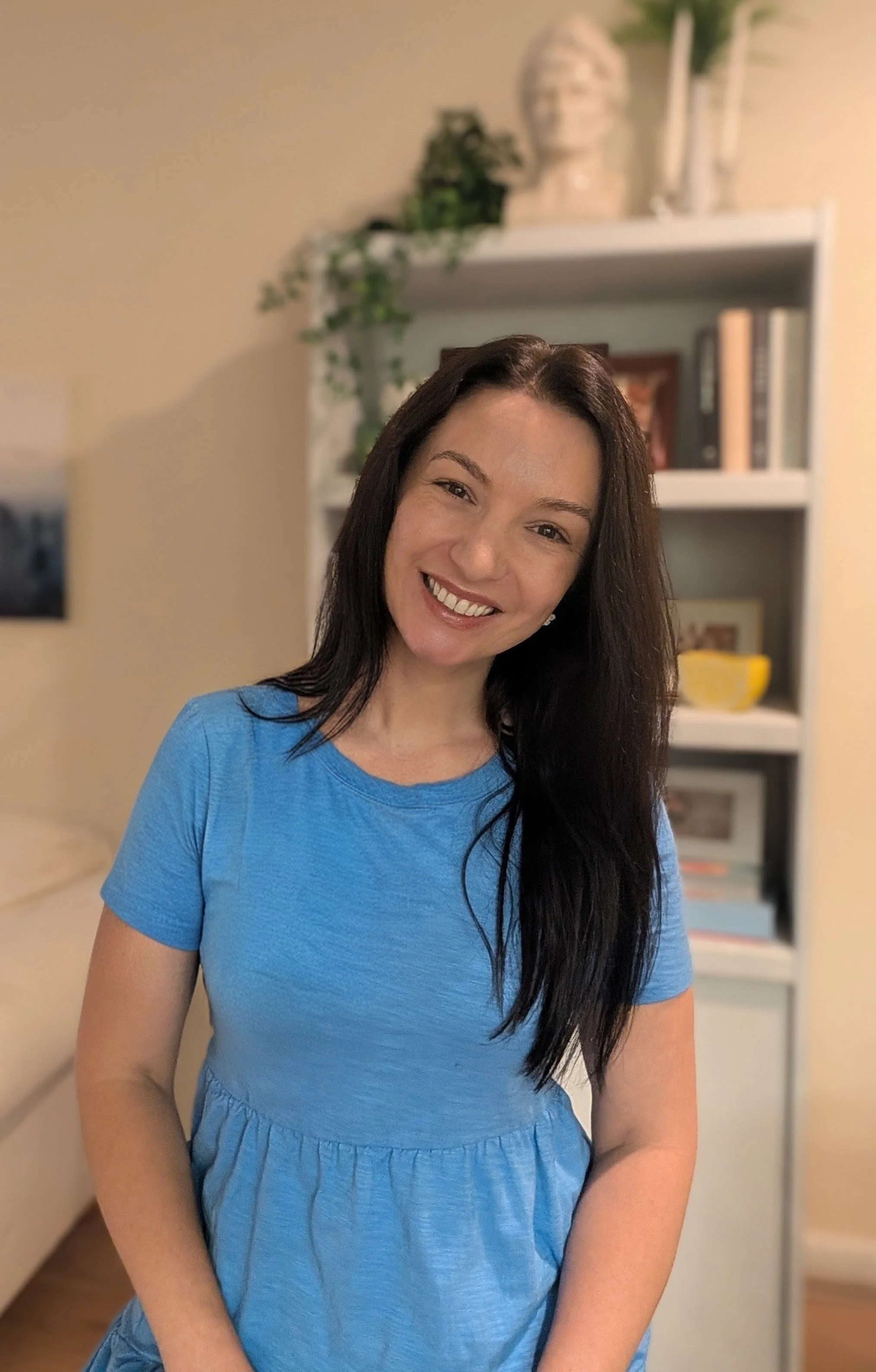 A woman with long dark hair smiling in a blue dress standing in a room with a bookshelf and a decorative head sculpture behind her.