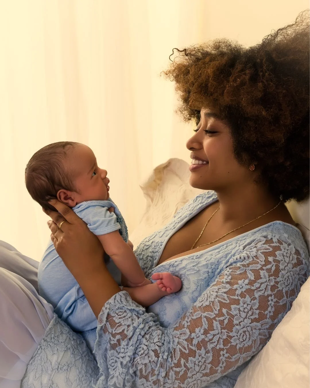 A woman with curly hair holding a baby in a blue outfit, looking at each other affectionately.
