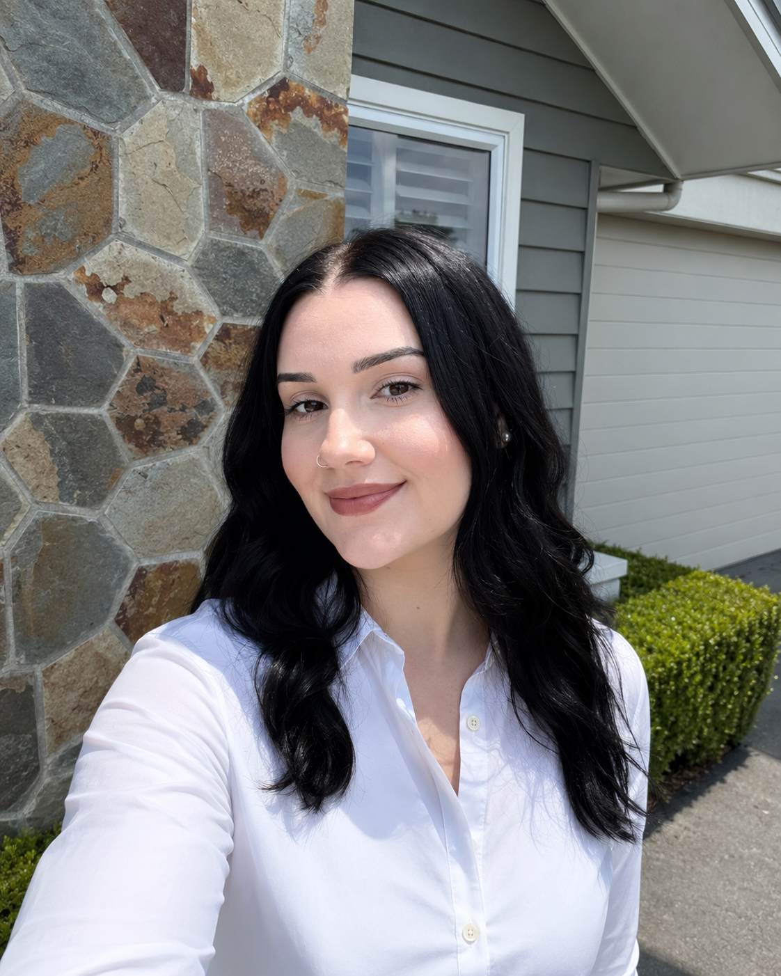 A young woman with black wavy hair, wearing a white button-up shirt, taking a selfie outdoors in front of a stone wall and a house with gray siding and a window.