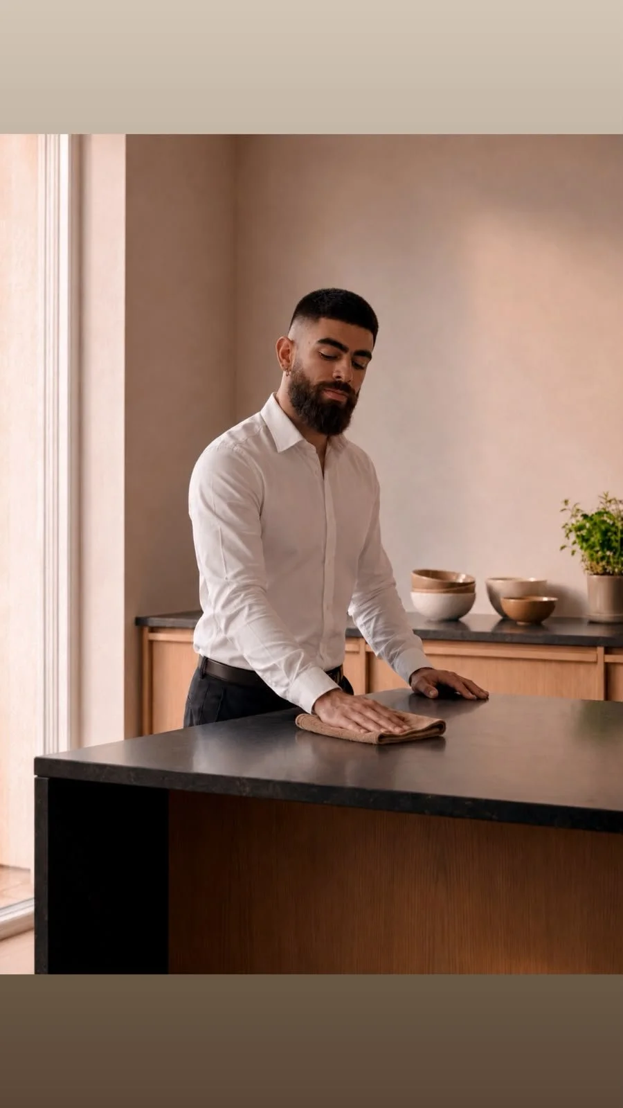 A man with a beard and wearing a white shirt wipes a kitchen counter with a towel.