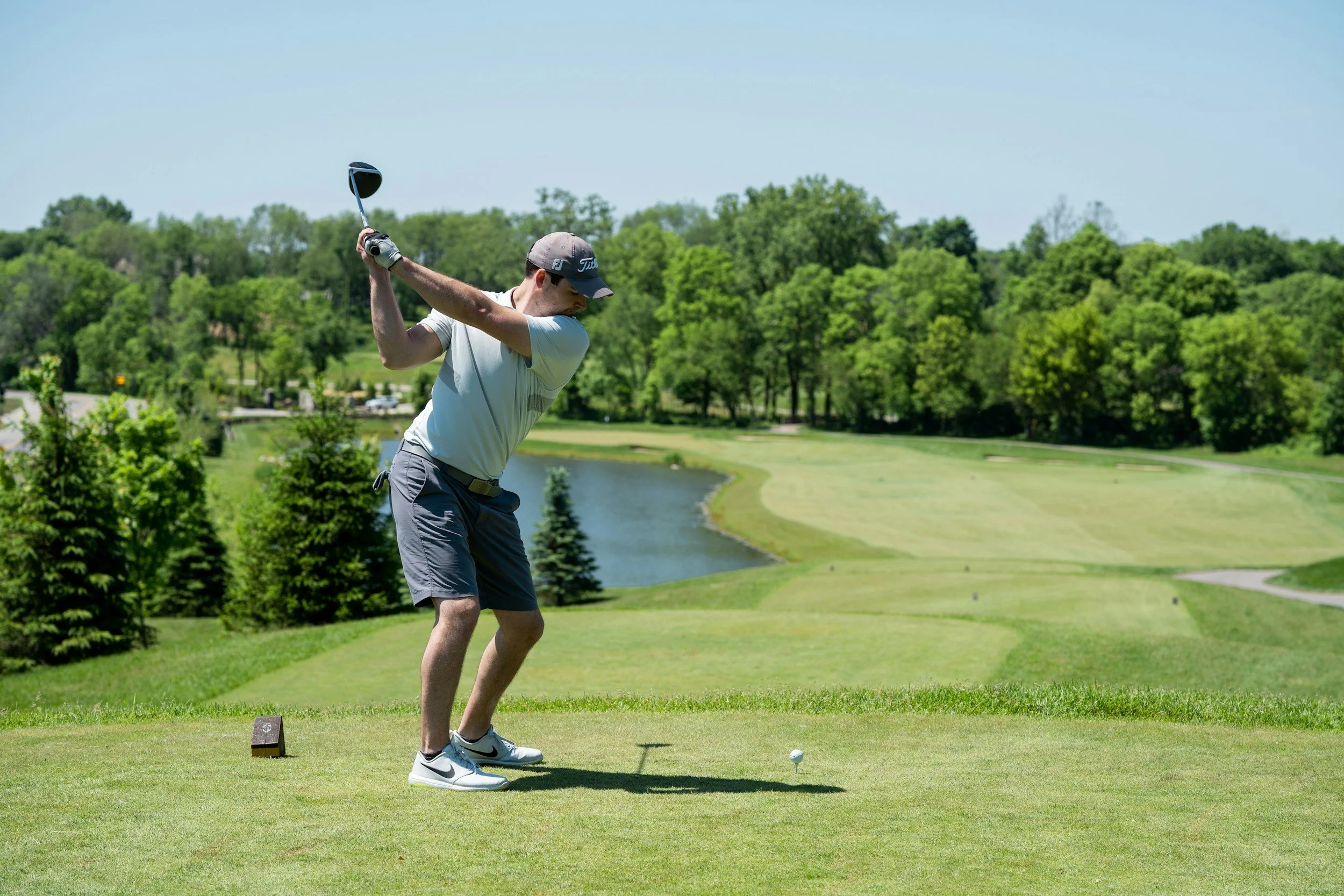 A man preparing to hit a golf ball on a green fairway near a water hazard, surrounded by trees under a clear blue sky.