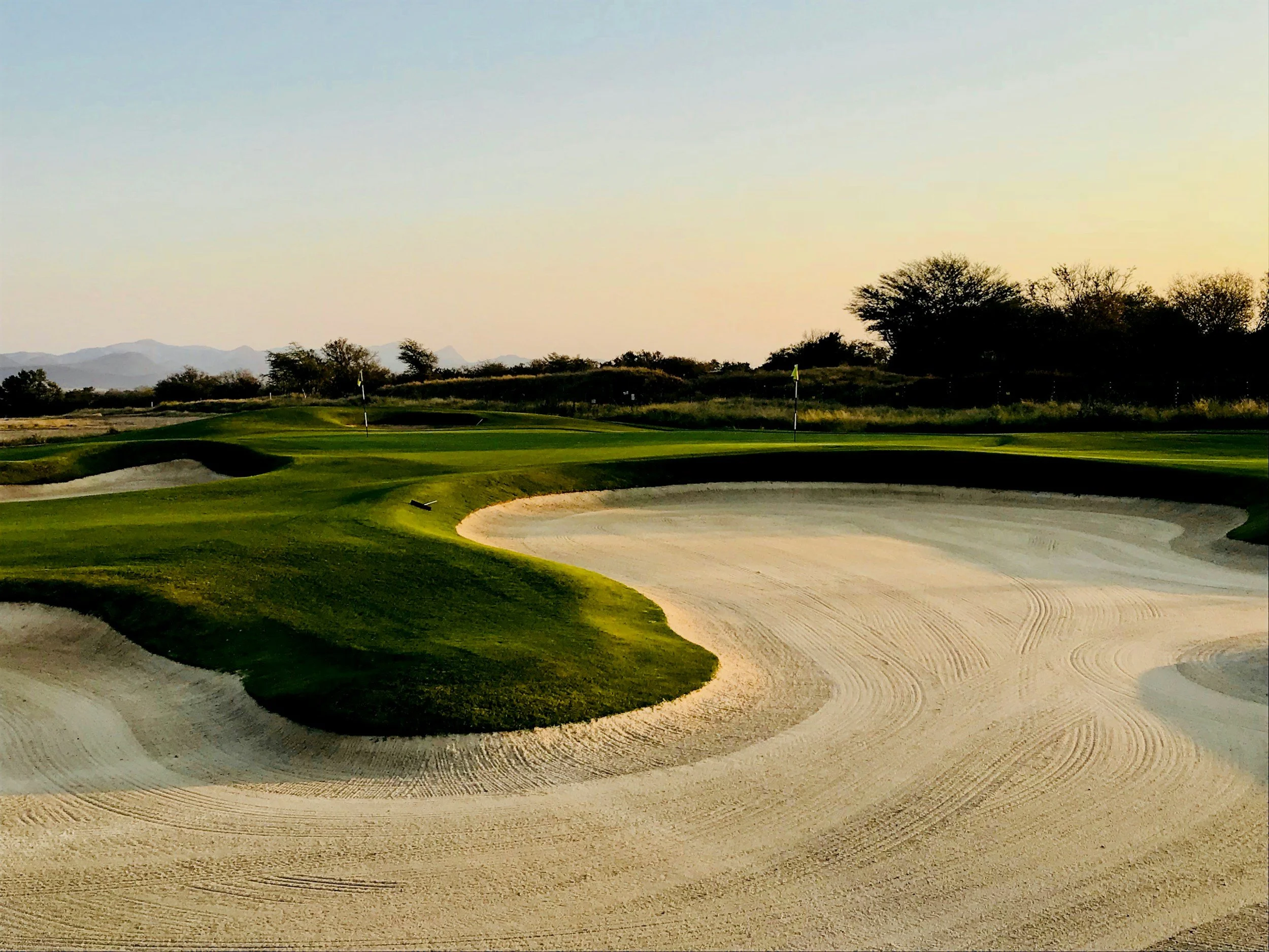 A golf course with sand bunkers around the green, some trees and hills in the background, and a clear sky.