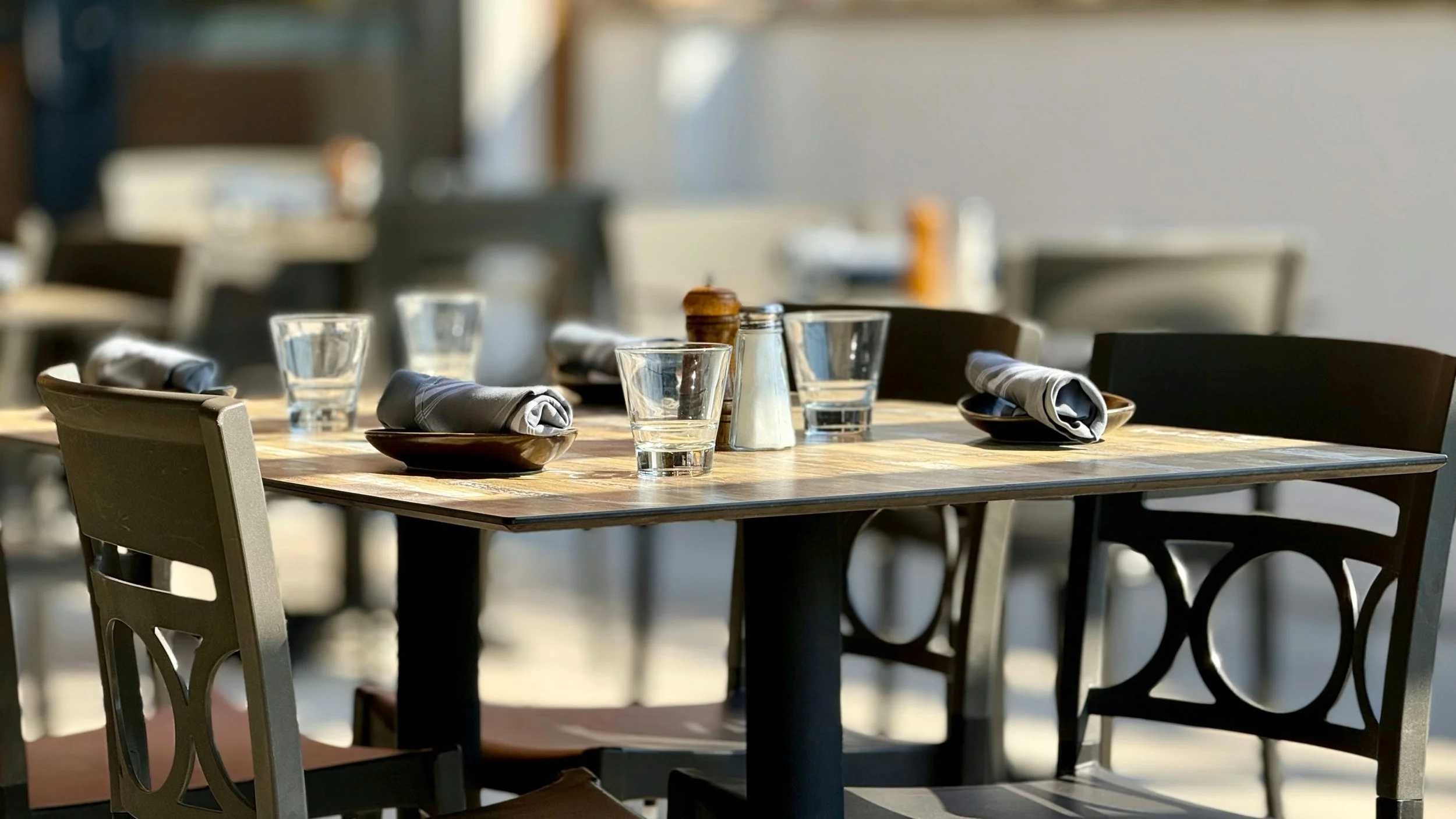 A wooden dining table set with glasses, napkins, plates, salt and pepper shakers, and chairs, in a bright restaurant setting.