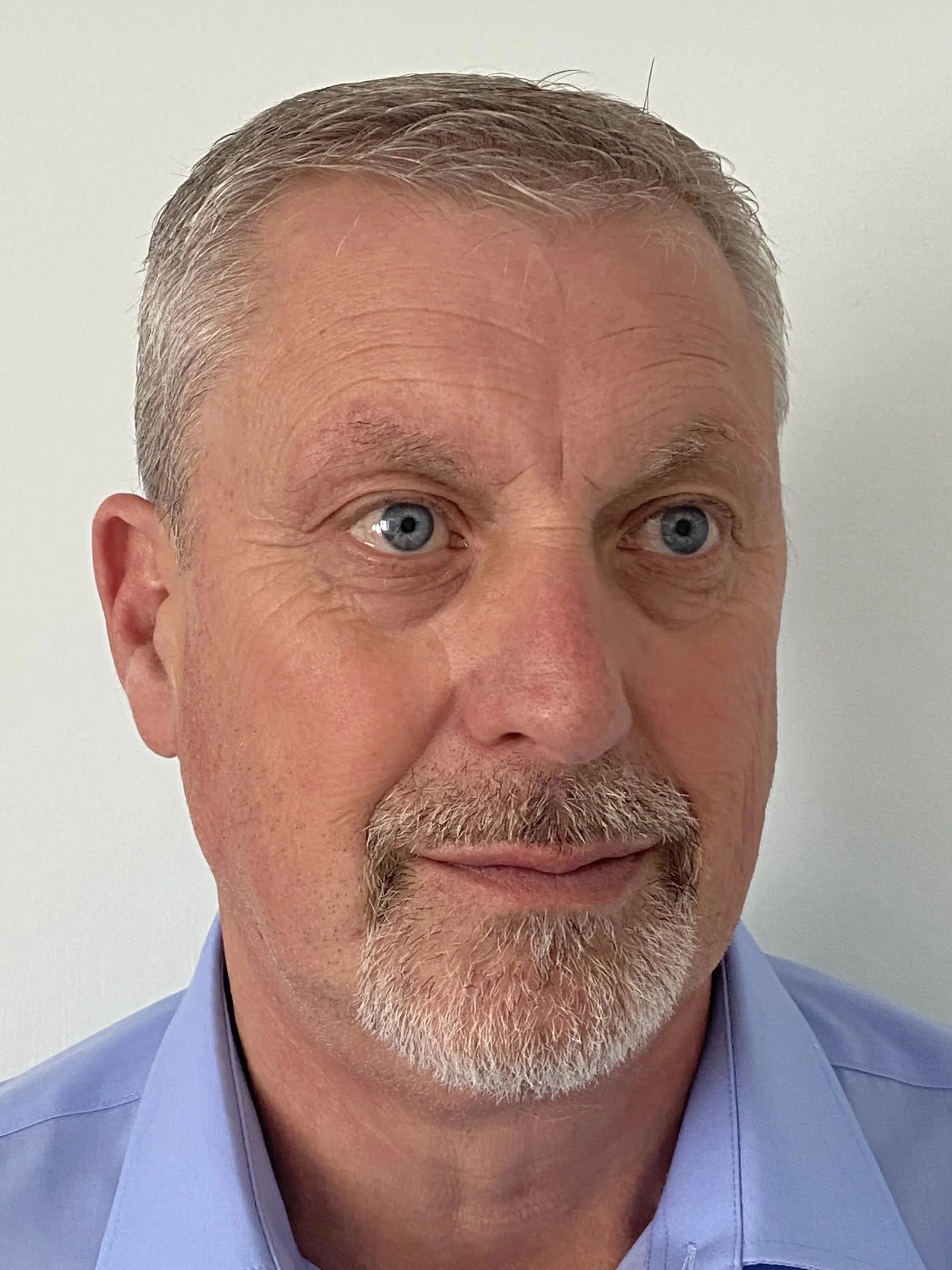 Close-up of a middle-aged man with blue eyes, short gray hair, and a beard, wearing a light blue collared shirt against a white background.