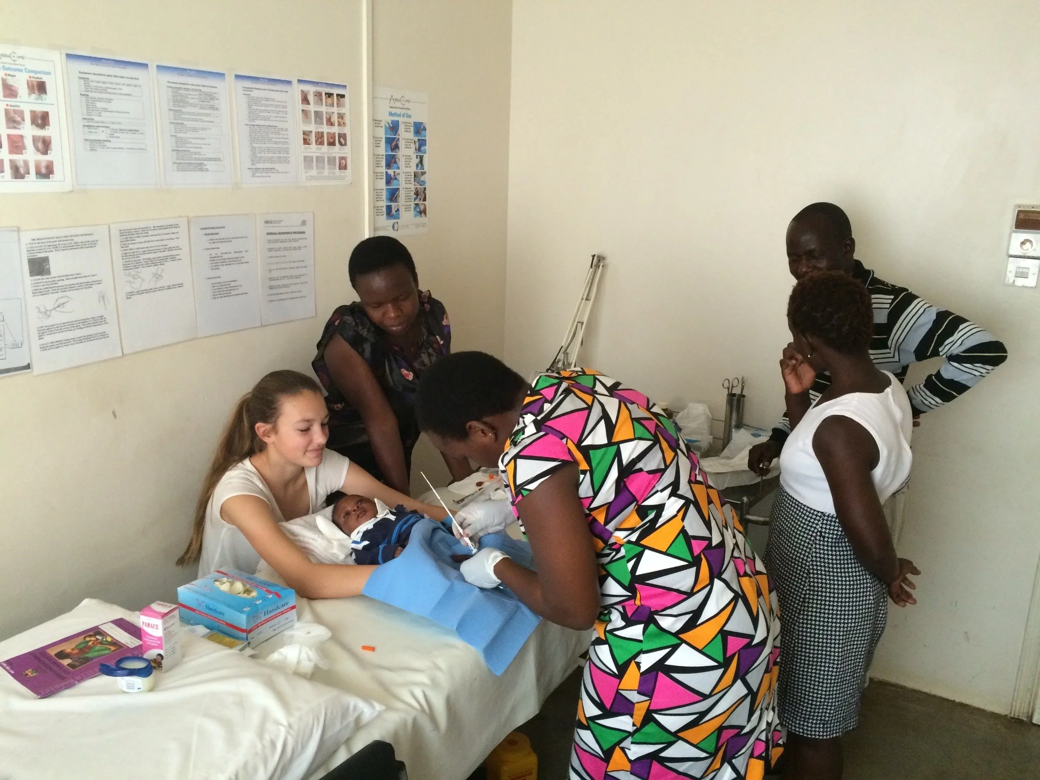 A woman in a colorful geometric dress attends to a baby lying on a hospital bed, with medical supplies nearby
