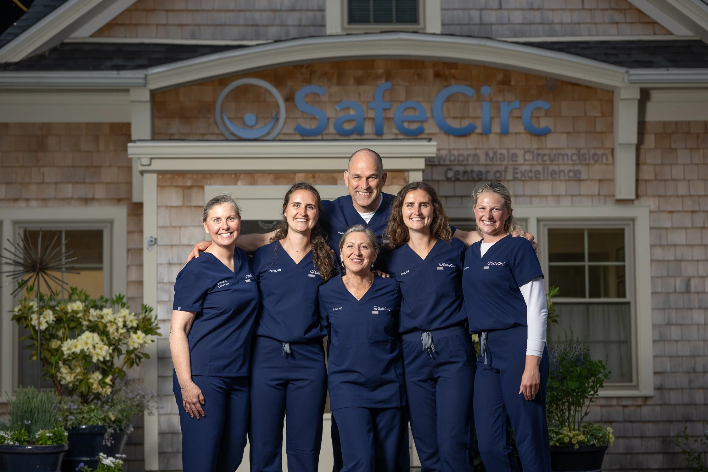 Group of six smiling healthcare professionals wearing navy blue scrubs standing in front of a building