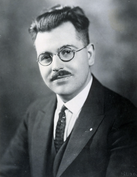 Black and white photo of a man with glasses, mustache, and neatly combed hair, wearing a suit and tie.