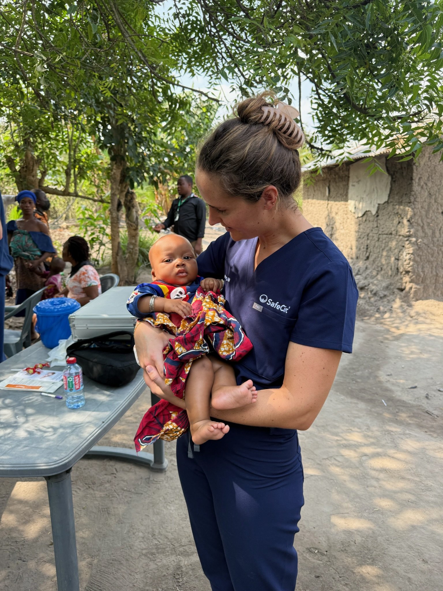 A woman in a navy blue uniform holding a baby girl dressed in colorful clothes at an outdoor setting with trees and a building in the background.
