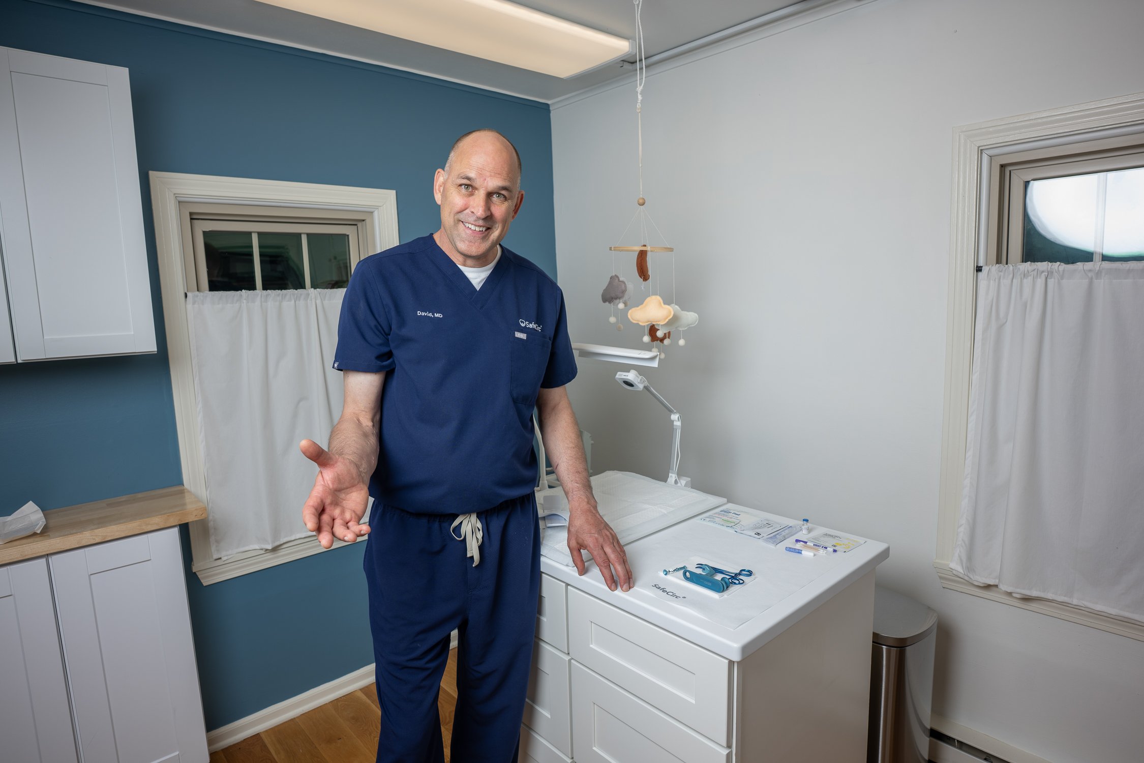 Dr David Tomlinson in blue scrubs standing in a medical examination room with a changing table, medical supplies, and a mobile toy