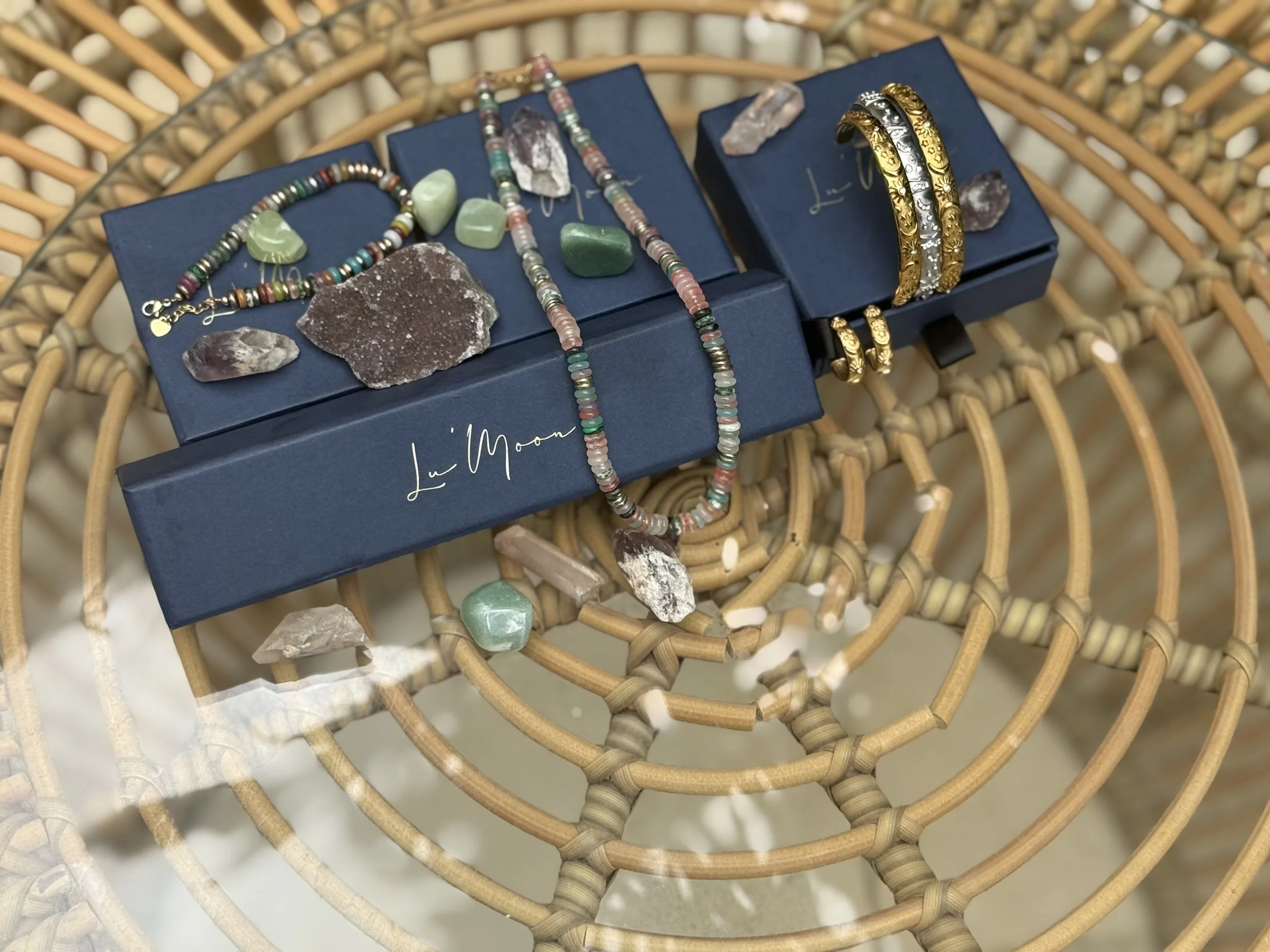 Various jewelry pieces, including bracelets, necklaces, and stones, displayed on blue boxes, placed on a woven rattan table.