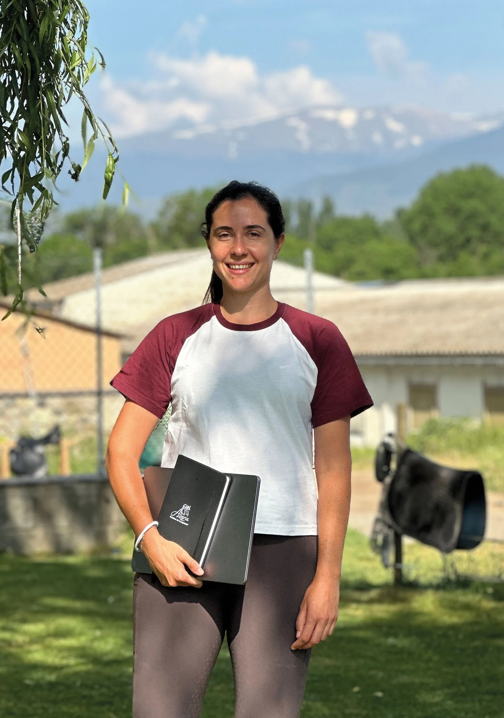 Ari Kairos con una camiseta blanca de Kairos Mountain Trail y pantalones gris oscuro, sosteniendo una libreta negra de Alegria, de pie en un área al aire libre con árboles y un fondo de montañas, bajo un cielo parcialmente nublado.