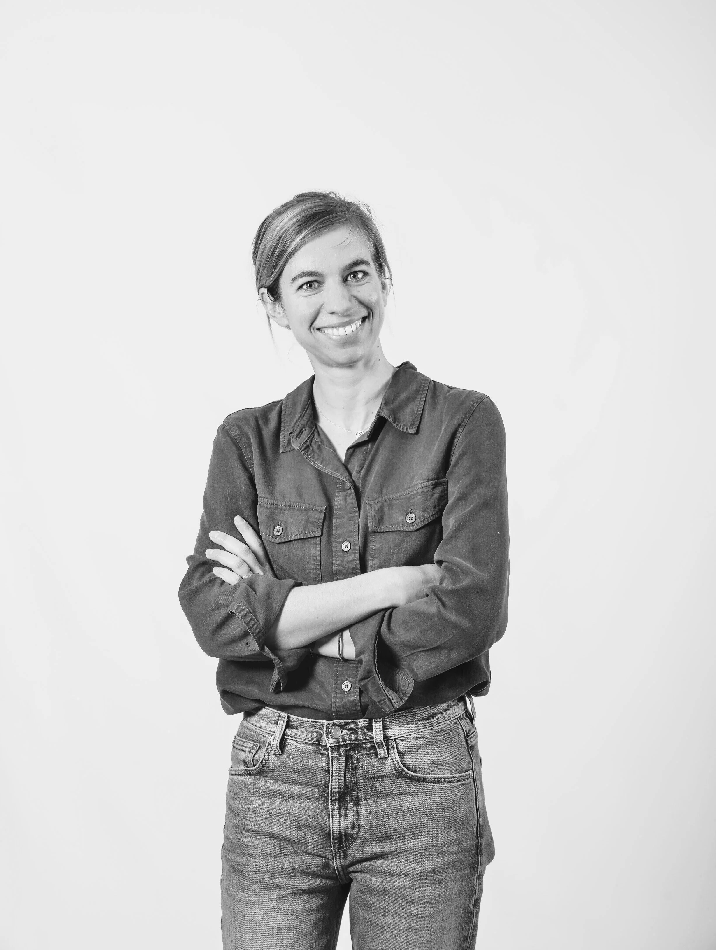 Une femme souriante, en chemise à manches longues, avec les bras croisés, pose contre un fond blanc.