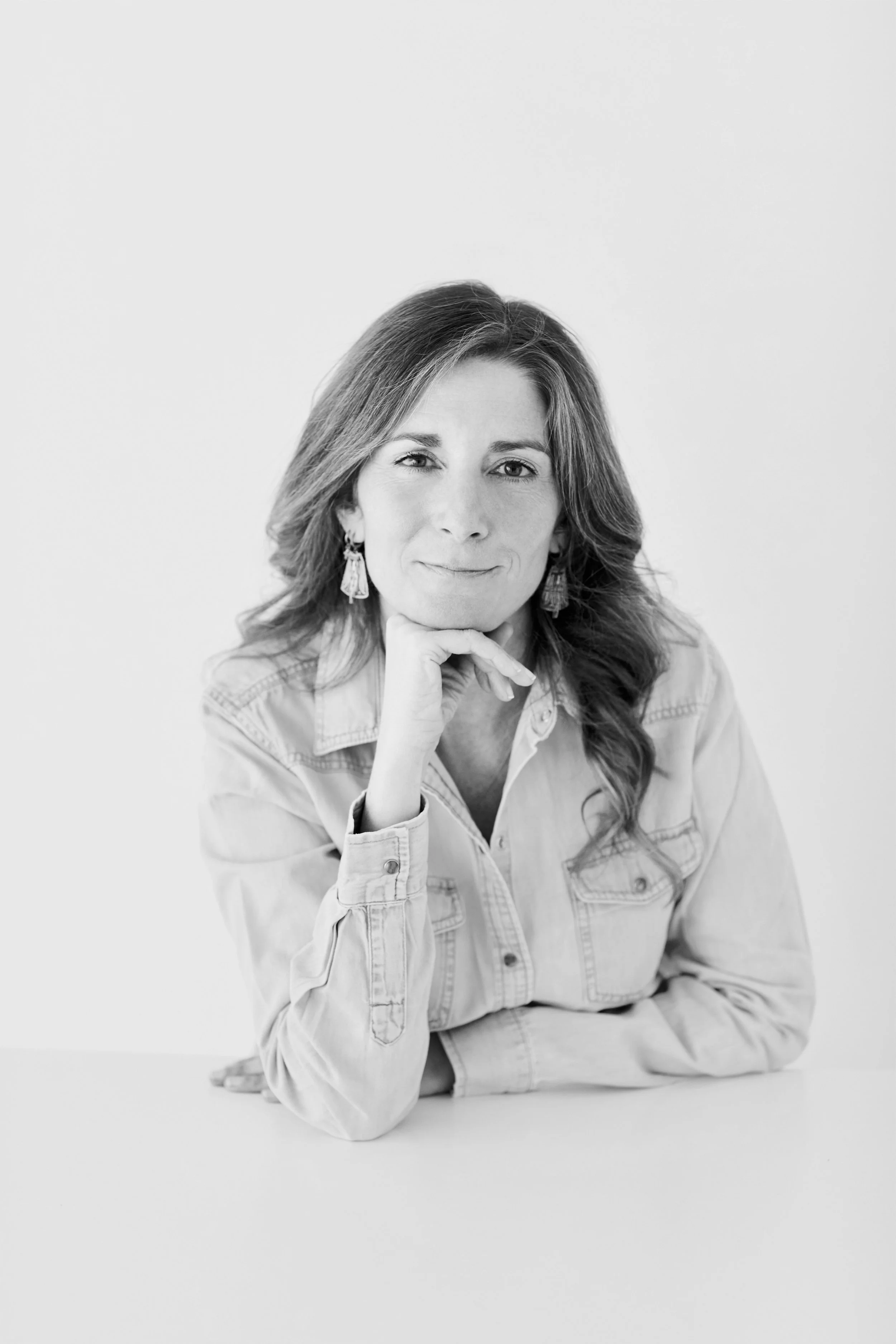 Portrait d'une femme souriante en noir et blanc, portant des boucles d'oreilles longues, avec une main sous le menton, vêtue d'une chemise en denim.