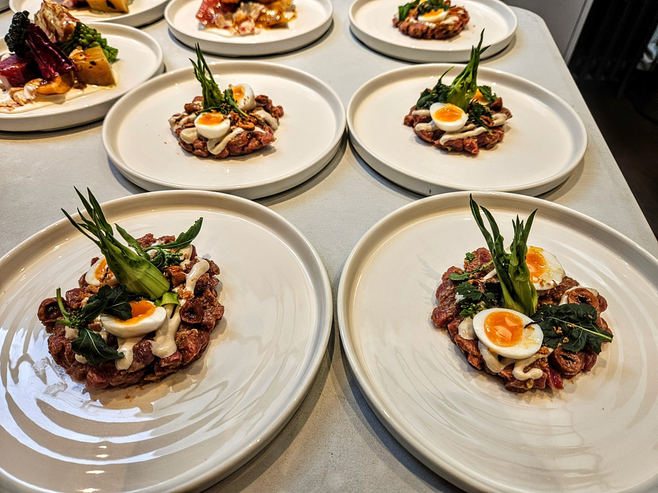 Plated dishes of steak tartare topped with soft-boiled eggs, garnished with green herbs, on white plates.