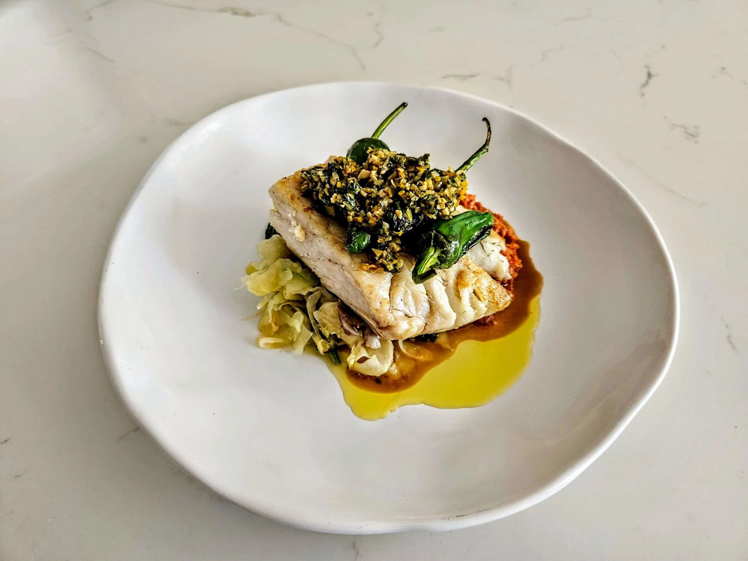 A plated dish featuring a piece of white fish topped with chopped herbs and vegetables, served over a bed of shredded vegetables with sauce and oil drizzle, on a white plate.