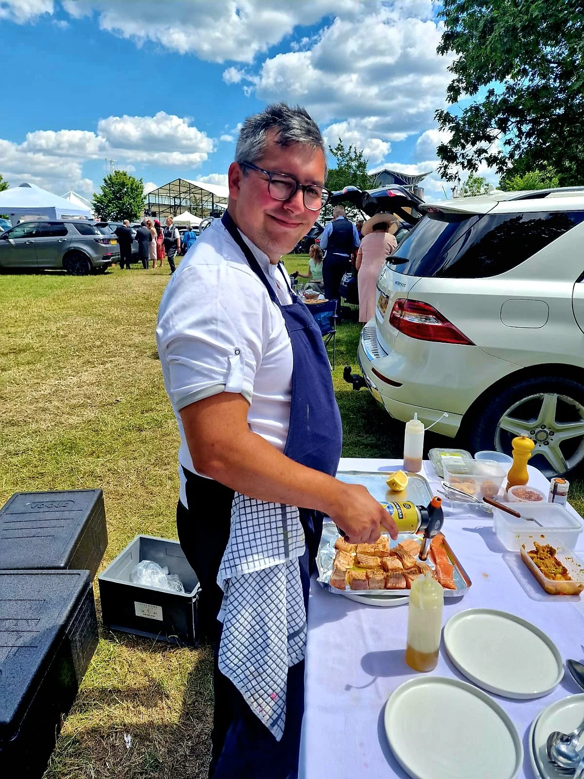 A man with glasses and gray hair wearing a white shirt and blue apron is preparing food at an outdoor event, standing next to a table with various ingredients and plates, with cars and tents in the background under a partly cloudy sky.