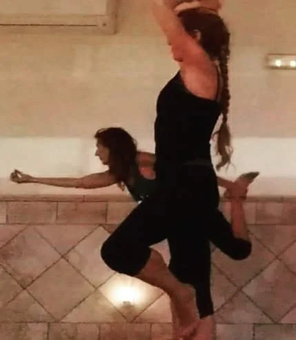 Two women practicing yoga or stretching together in a studio with tiled walls and a mirror.
