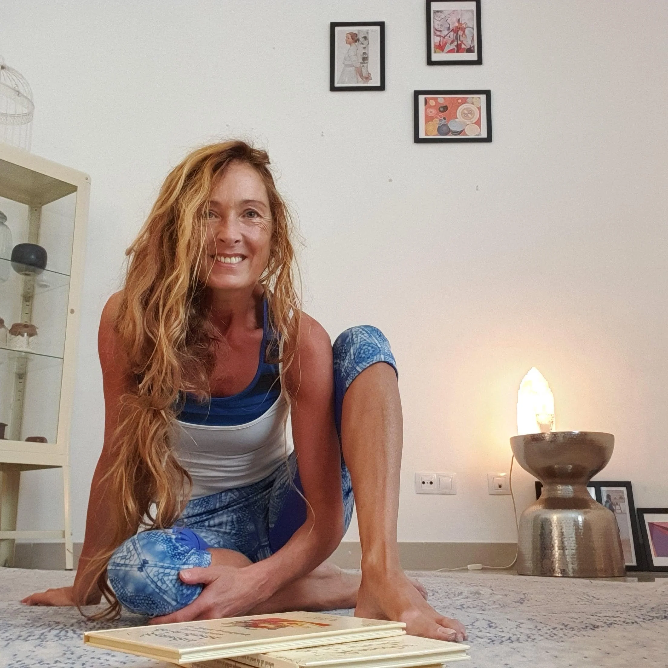 A woman with long red hair smiling while lying on the carpeted floor, reading a book. Another person with blue patterned clothing is partly visible behind her. The room has white walls decorated with three framed pictures and a white bookshelf on the left. There is a table or stool with a tall lamp and a large glass jar on the right.