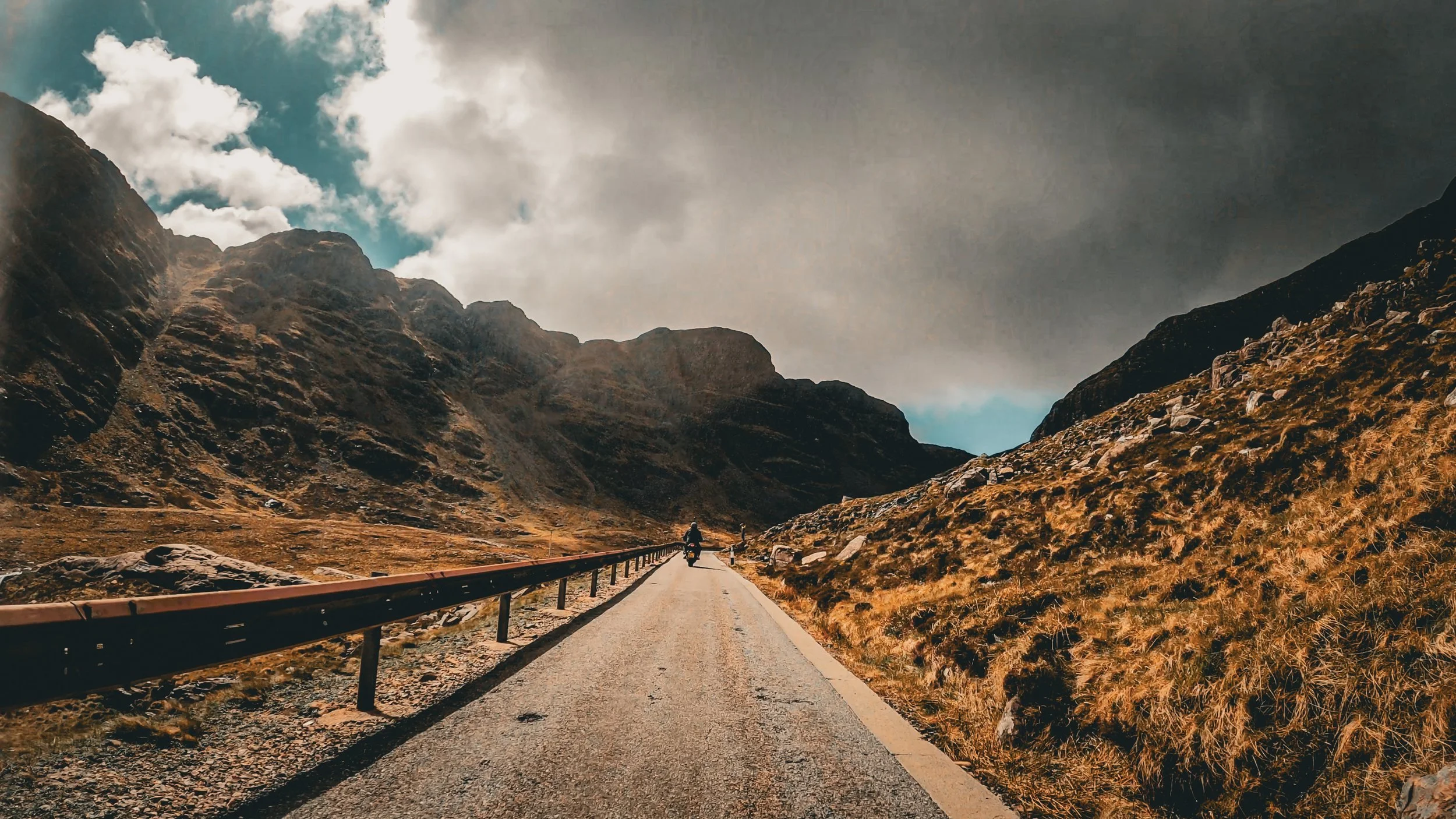 A motorcyclist riding on a narrow mountain road surrounded by rugged hills with a dramatic cloudy sky overhead.