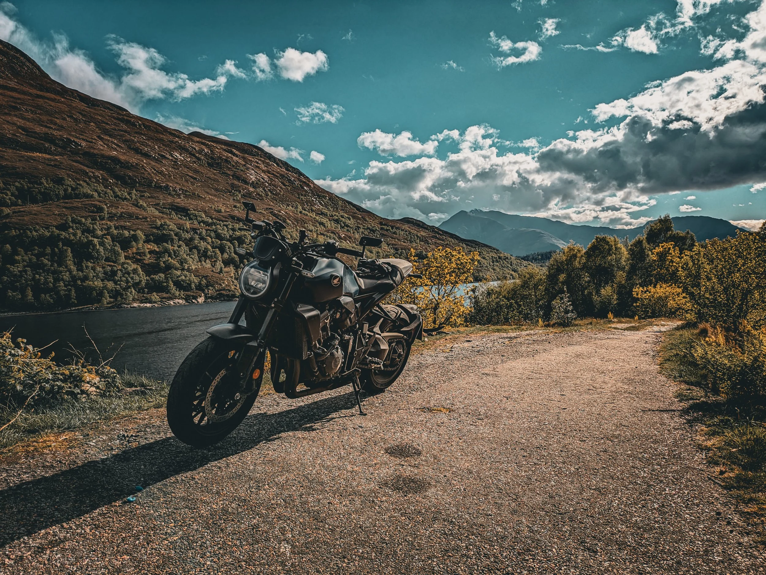A black motorcycle parked next to a body of water on a gravel road surrounded by trees and mountains under a partly cloudy sky.
