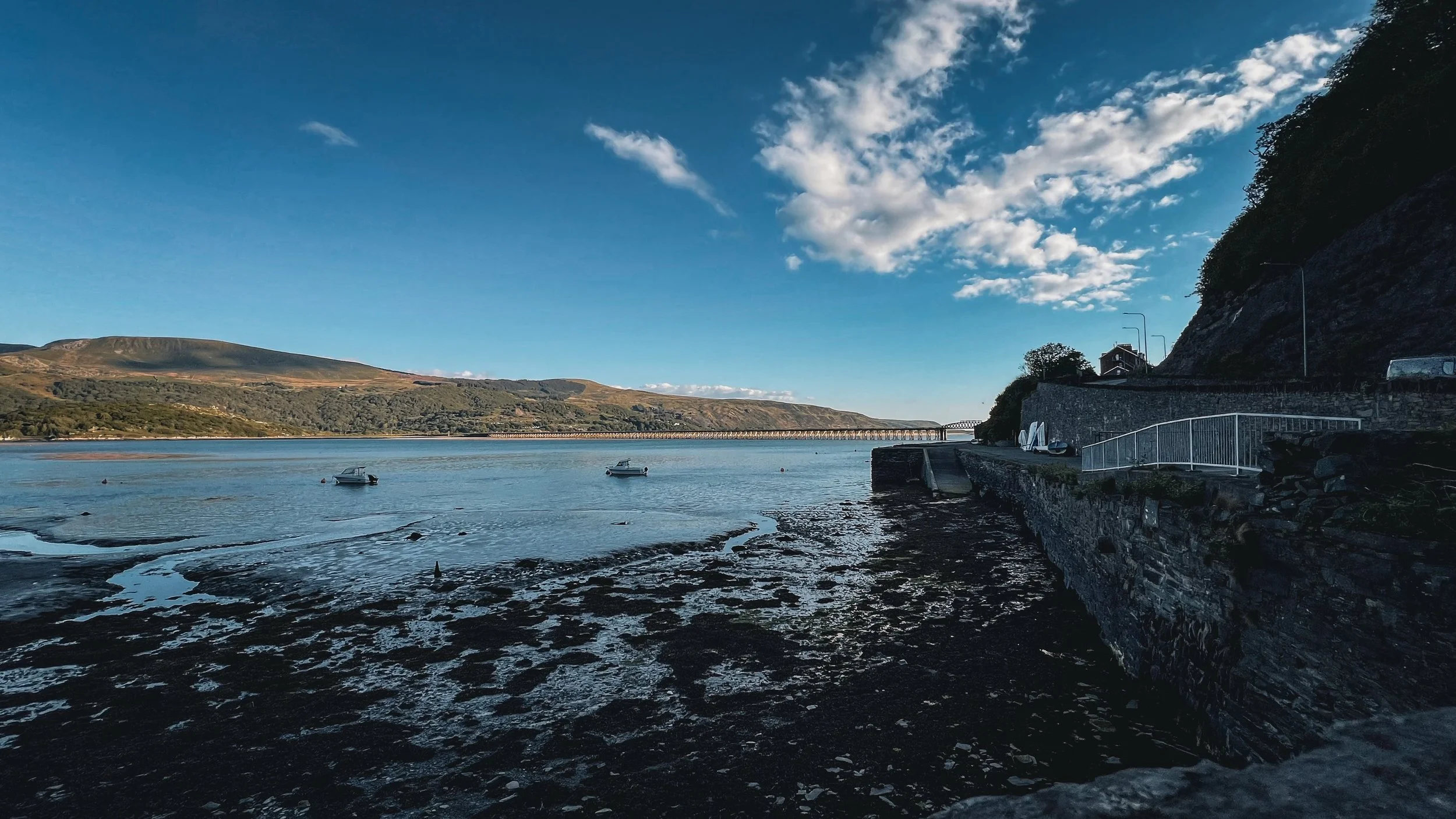 View of a calm river with small boats, surrounded by hills, with a stone wall along the shore and a scenic sky with clouds.