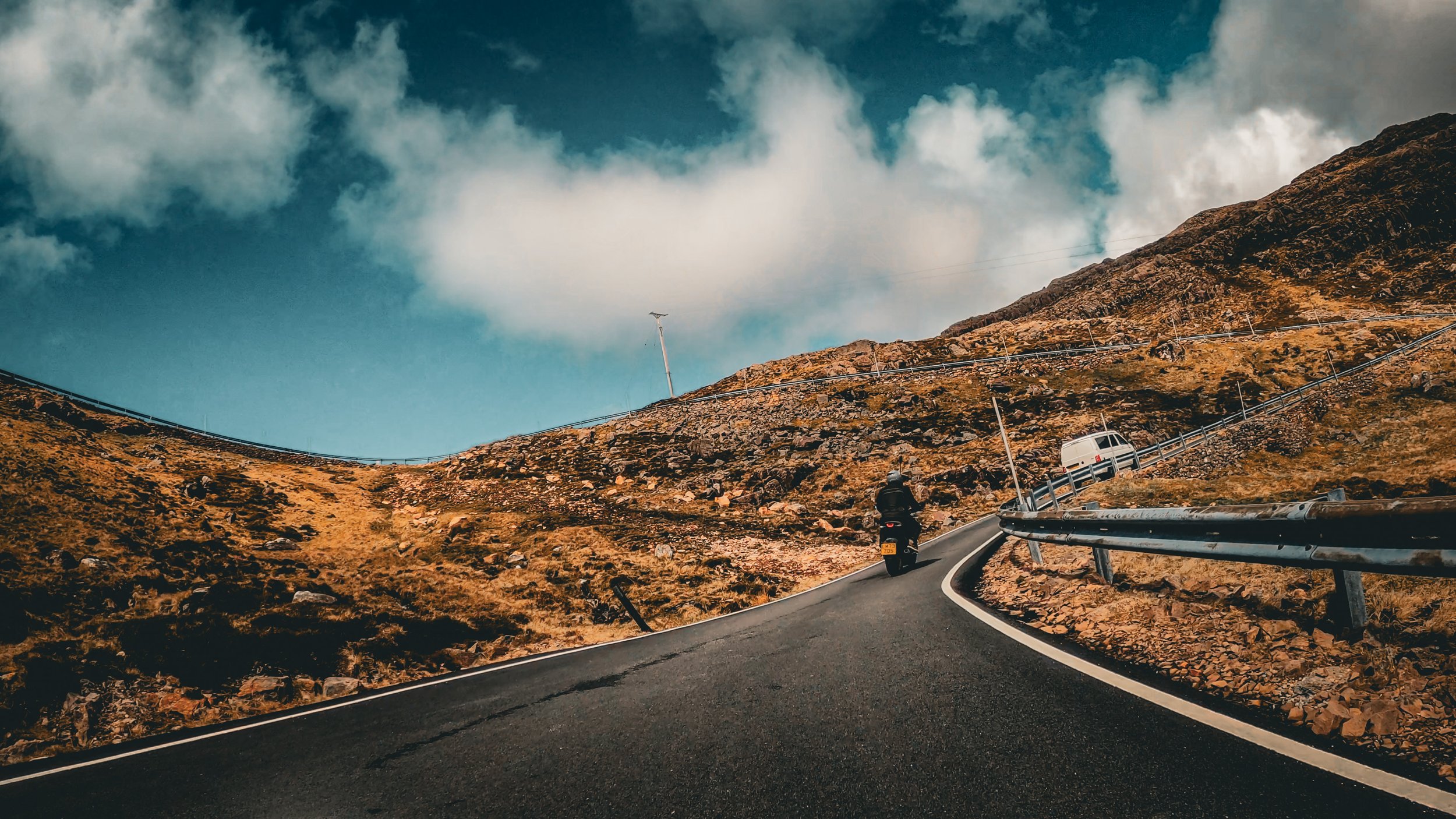 A winding mountain road with a motorcyclist and a car, rocky terrain, a wind turbine, and a partly cloudy sky.