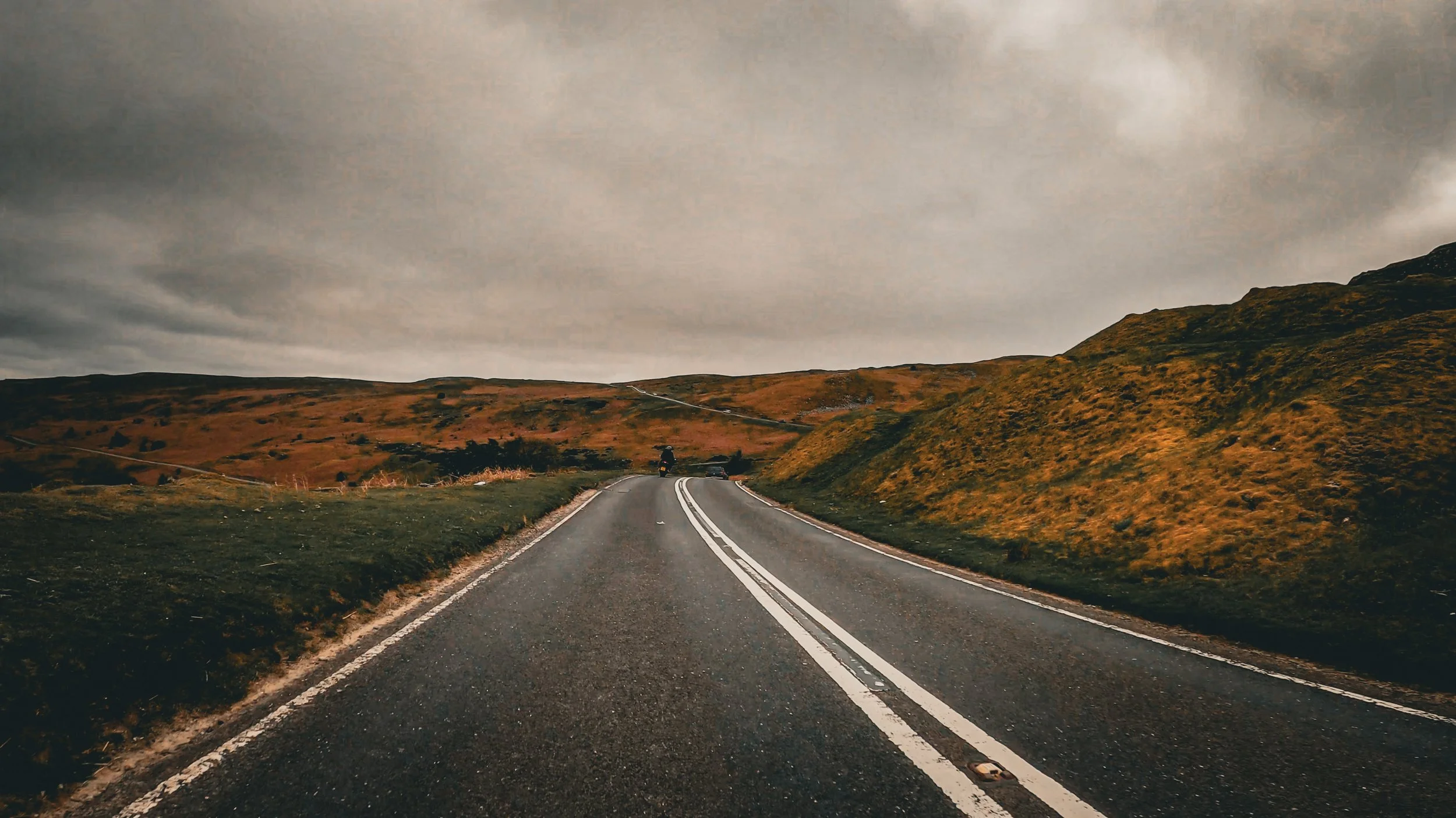 A winding two-lane road in a hilly landscape with grassy and rocky terrain under a cloudy sky.