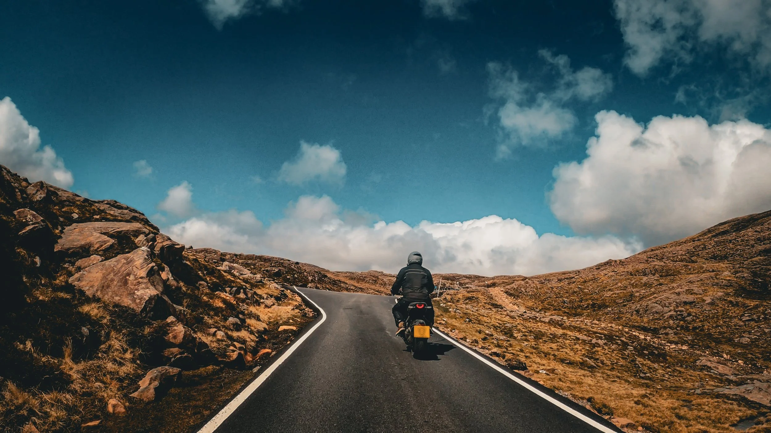 A person riding a motorcycle on a winding mountain road with rocky terrain and a blue sky with scattered clouds.