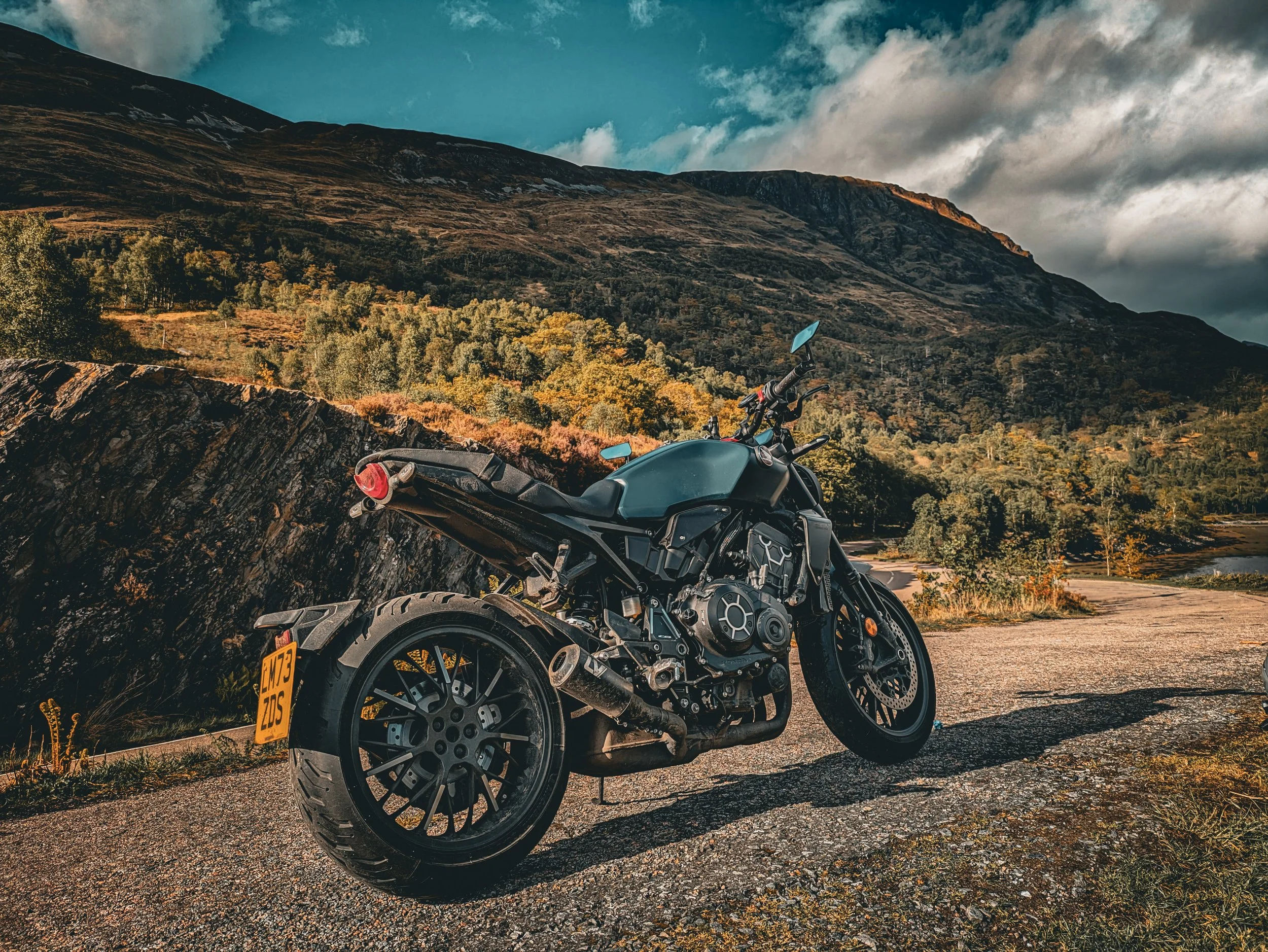 A black motorcycle parked on the side of a mountain road with a scenic view of mountains and trees in the background, under a partly cloudy sky.