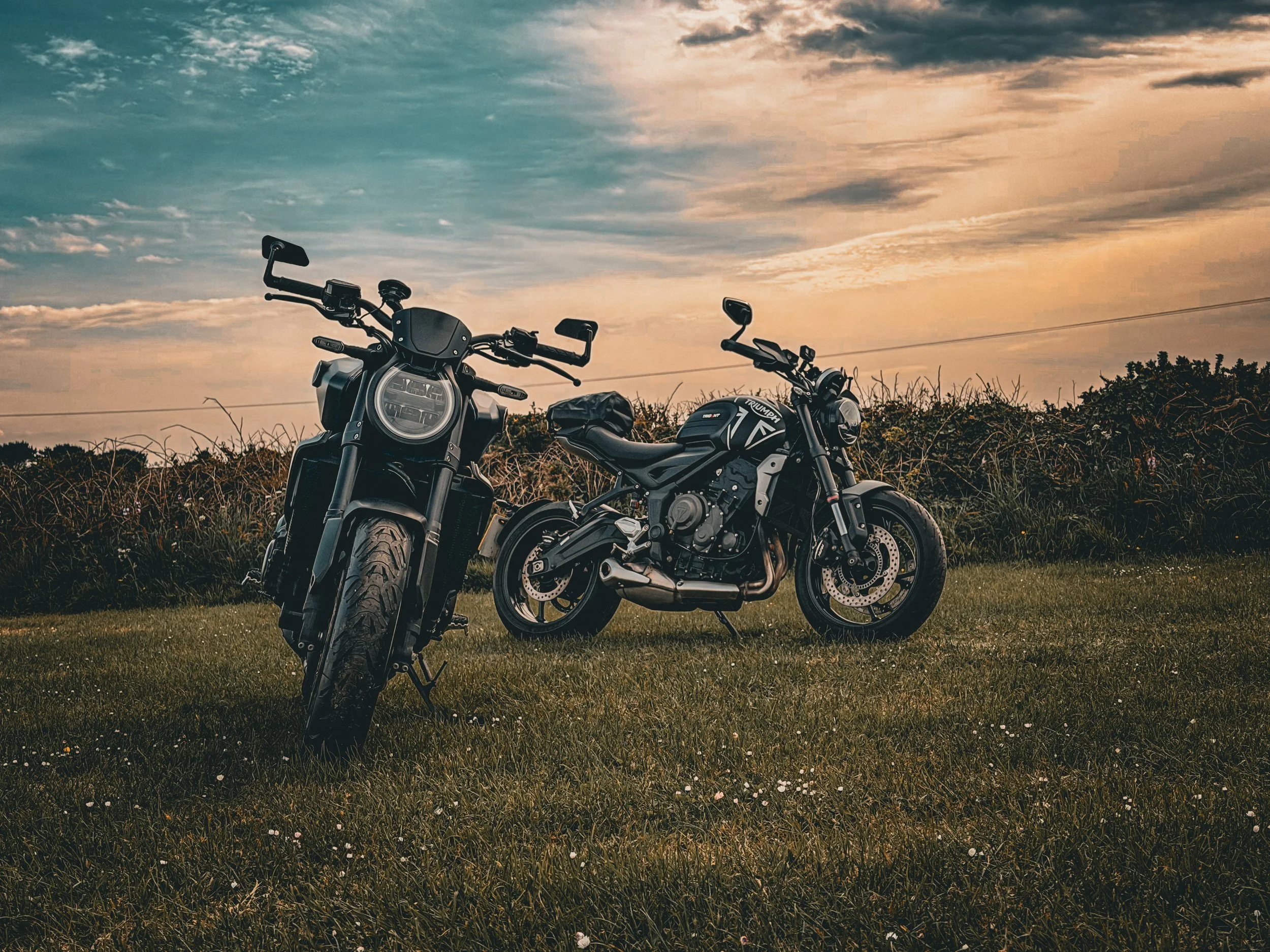 Two motorcycles parked on a grassy field during sunset or sunrise, with a sky filled with scattered clouds and a mix of blue and orange hues.