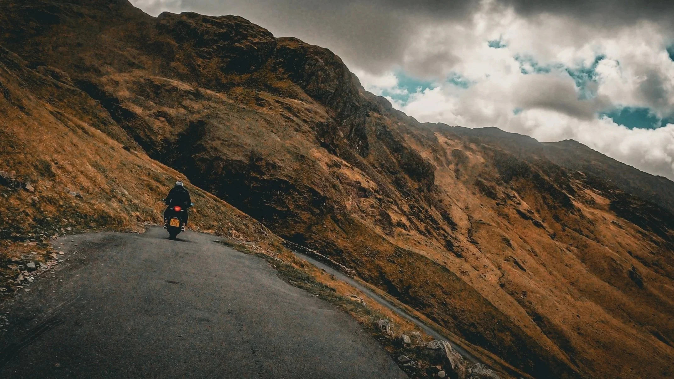Motorcycle rider traveling on a narrow mountain road surrounded by steep, rocky terrain with overcast sky.