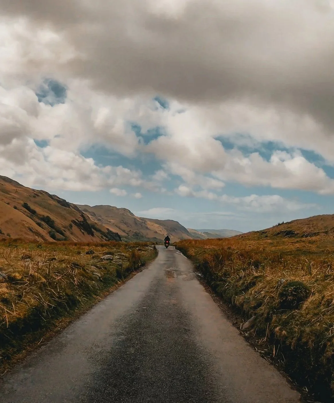 A lone motorcyclist riding down a narrow, paved country road surrounded by rolling hills and a cloudy sky.