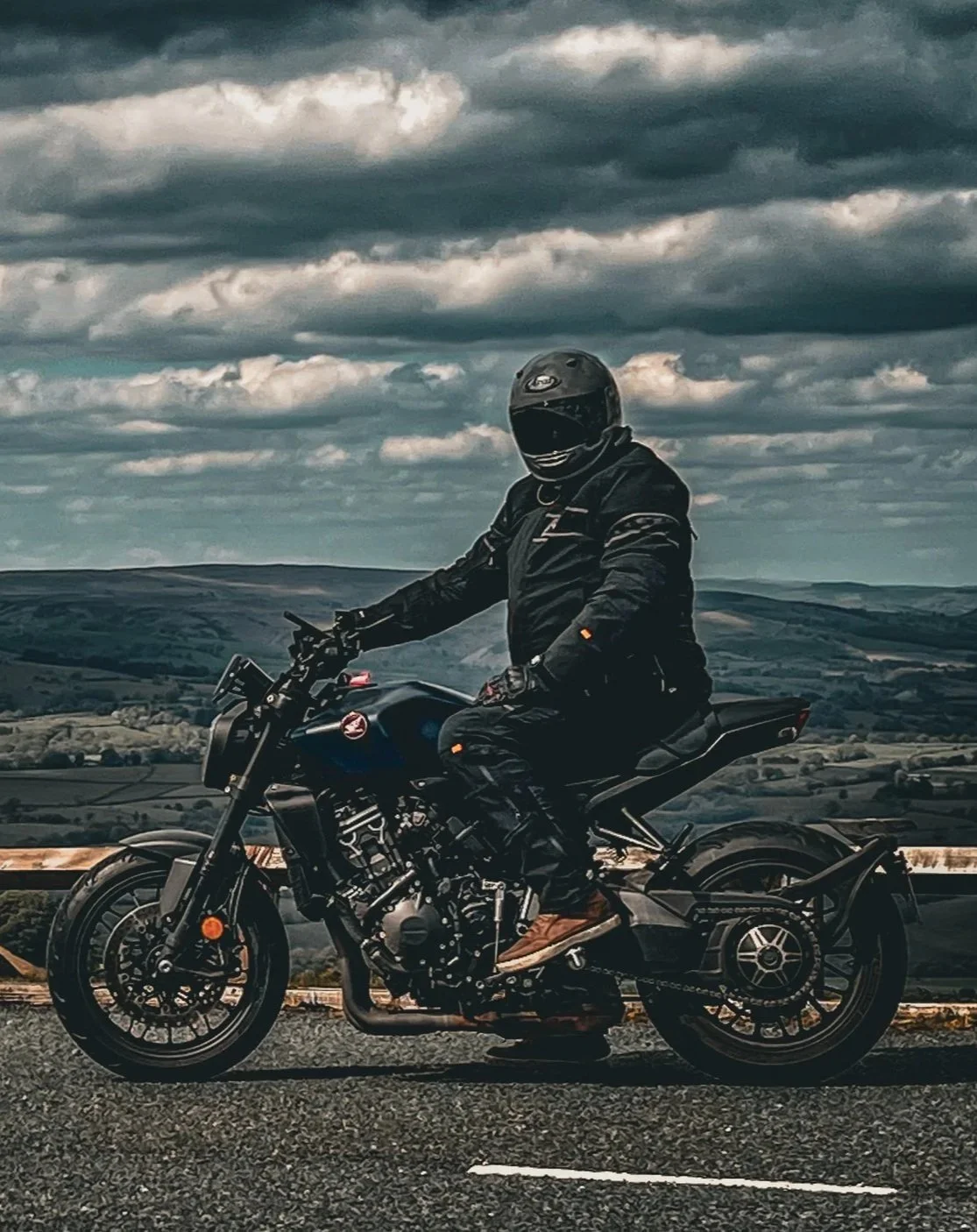 Motorcyclist wearing black gear and helmet riding a black motorcycle on a cliffside road with scenic landscape and cloudy sky in the background.