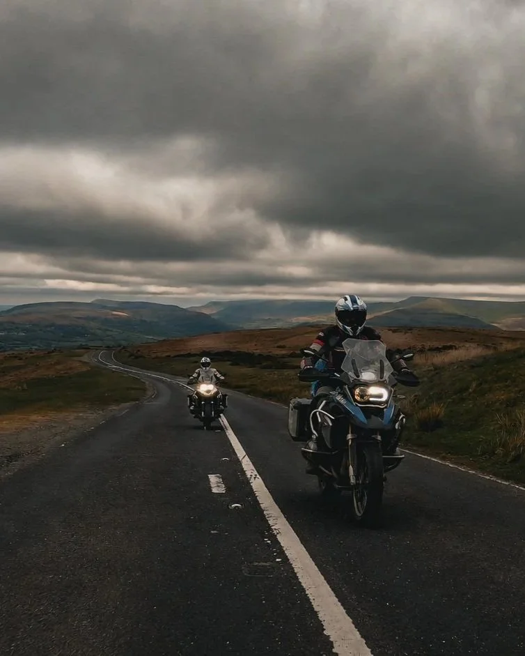 Two motorcyclists riding on a winding rural road under dark, cloudy sky with mountains in the background.
